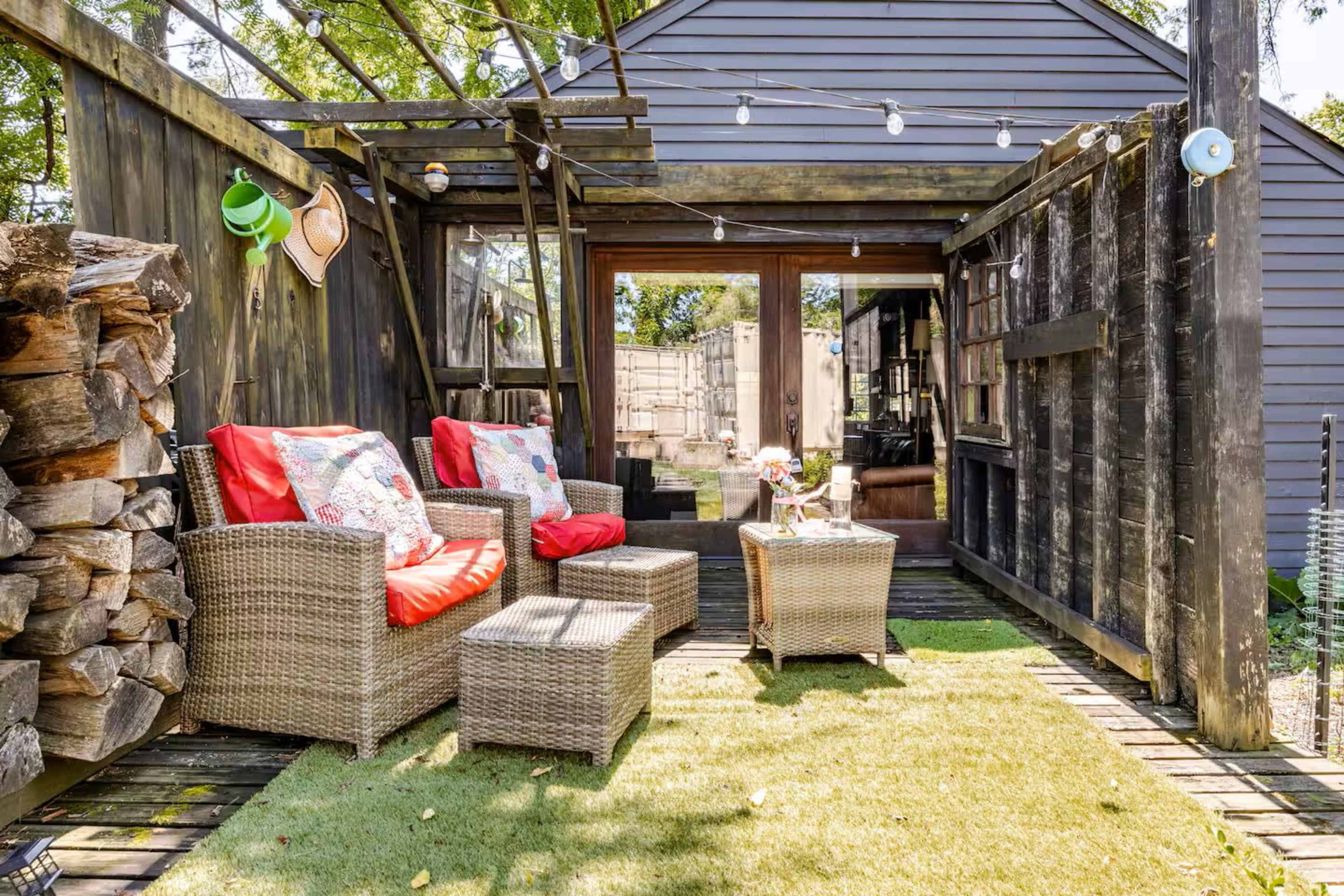The image shows a cozy outdoor seating area with two wicker chairs and a table, surrounded by wooden walls and a stack of firewood, beneath string lights and greenery.