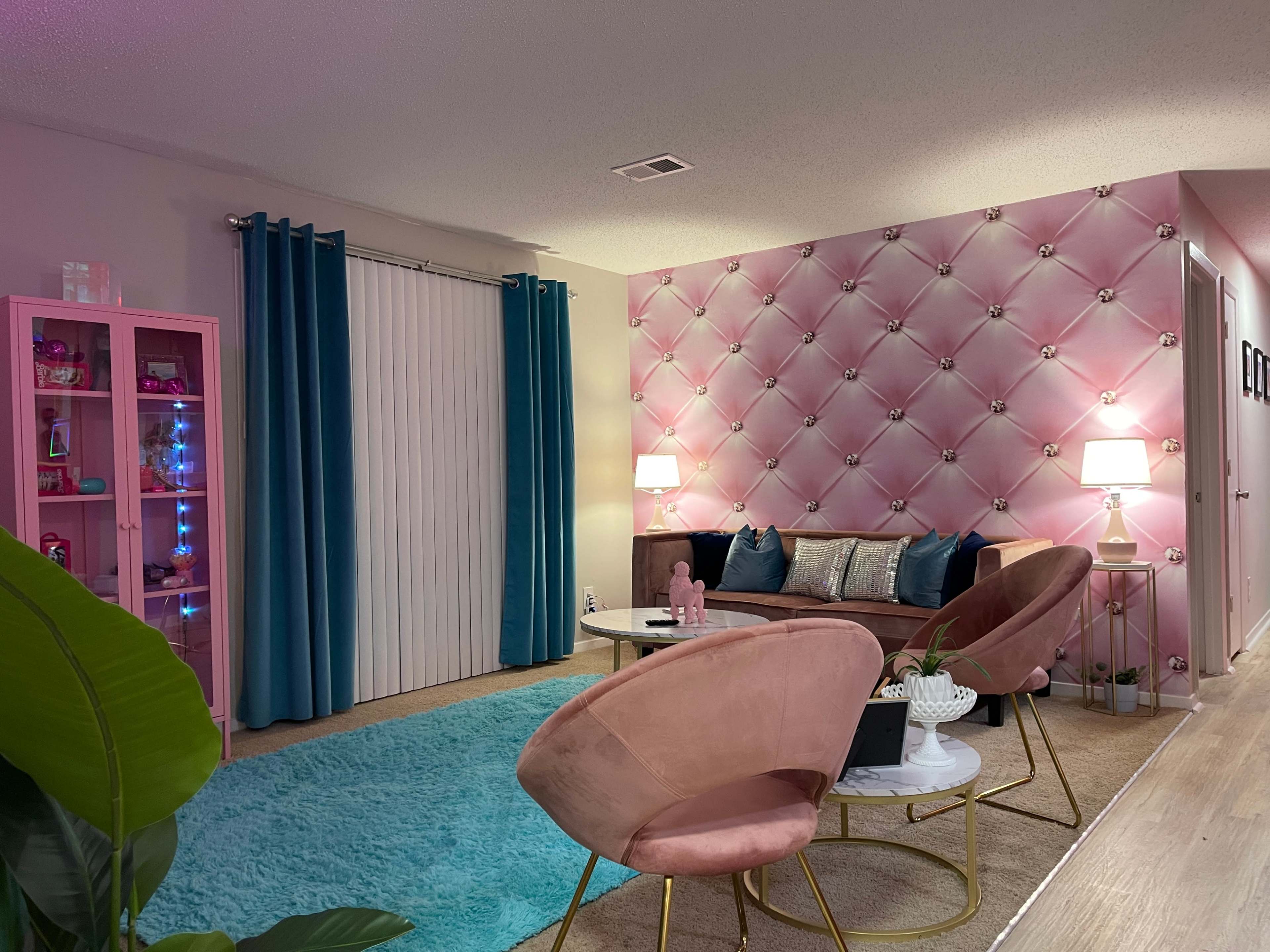 A modern living room featuring a pink tufted accent wall, teal curtains, a plush area rug, and stylish furniture including a sofa and two pink chairs.