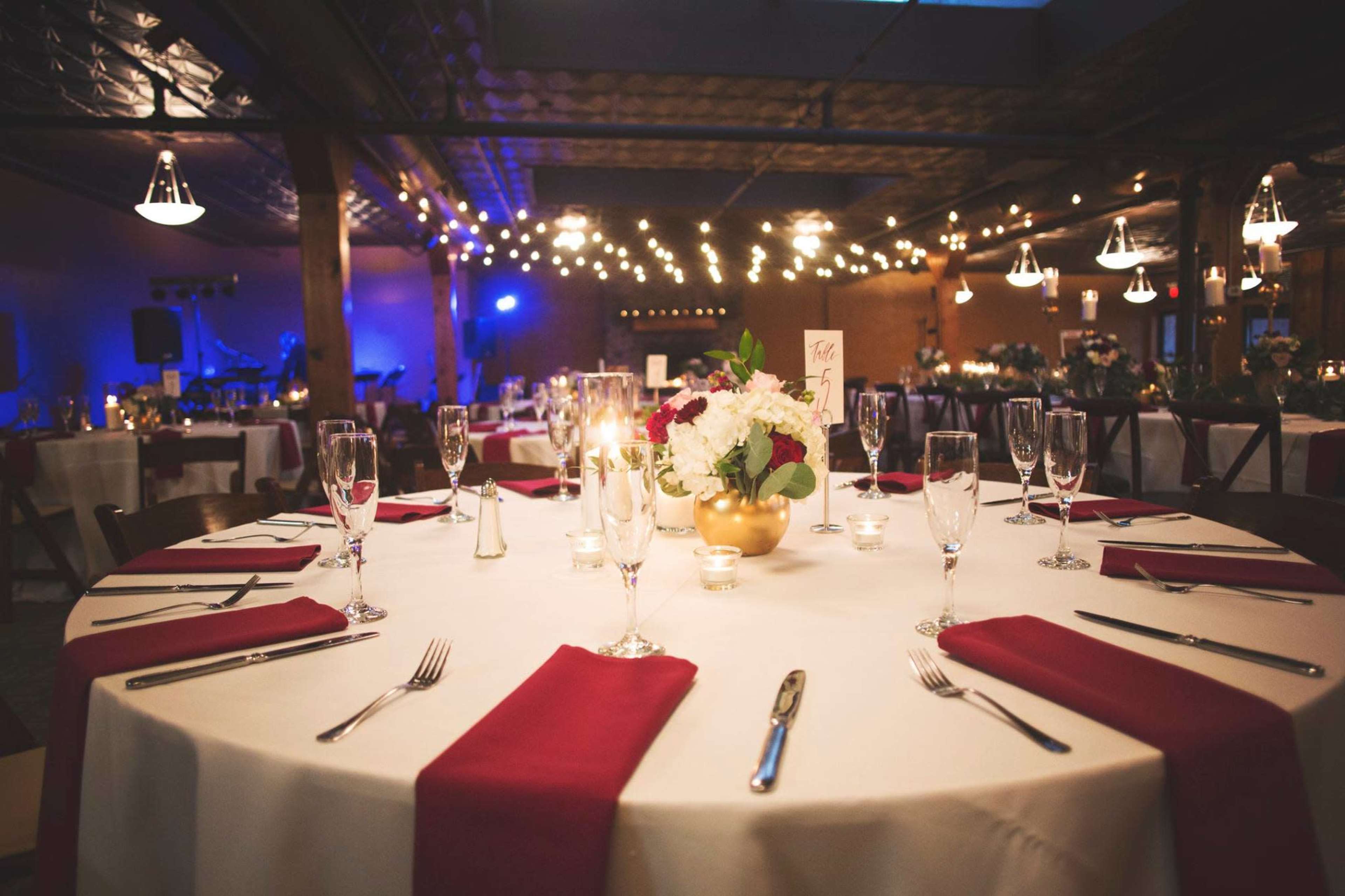 A round dining table is set with elegant tableware, red napkins, and a floral centerpiece, illuminated by ambient lighting and string lights above.