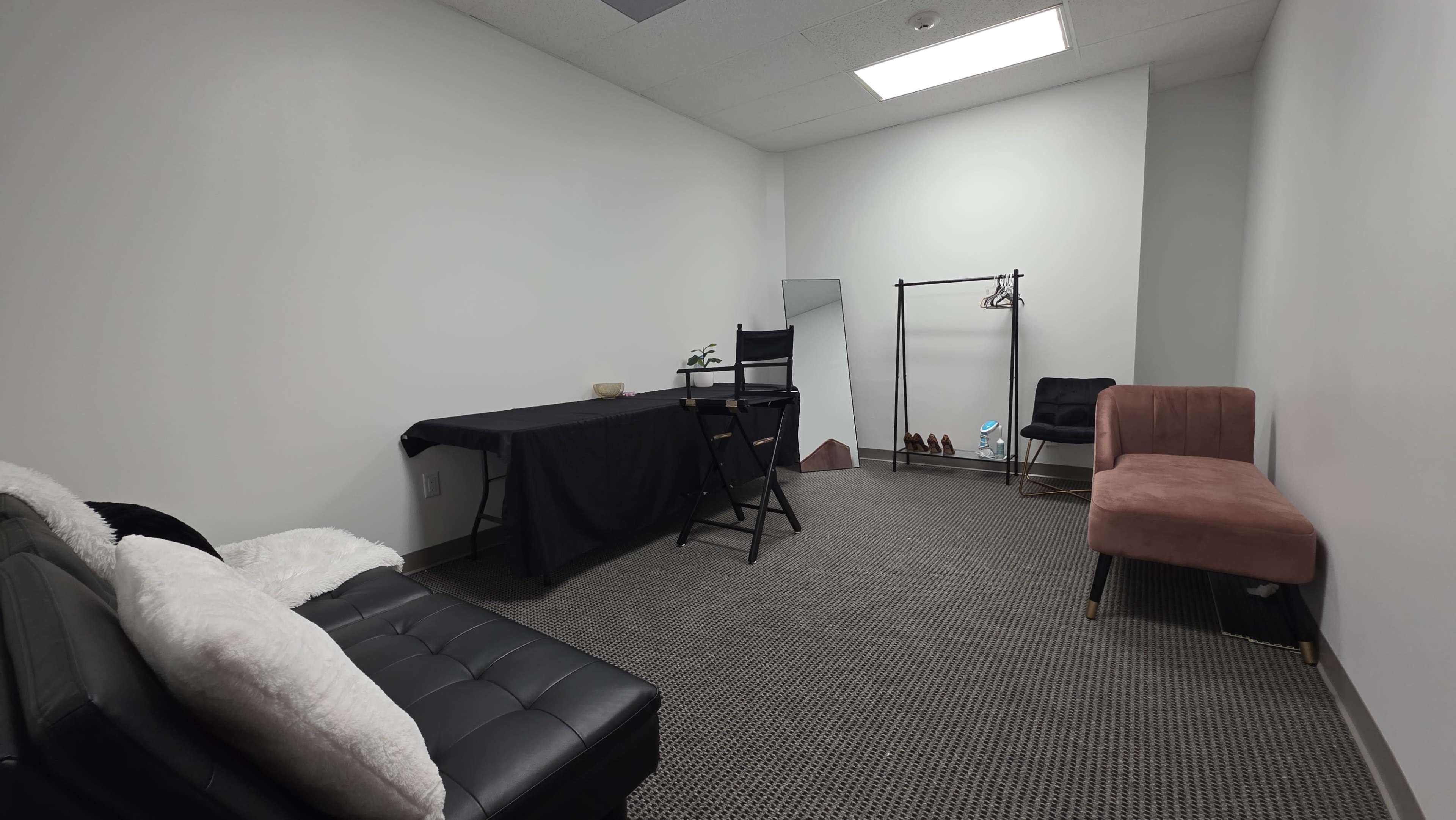 The image shows a small, empty room furnished with a black futon, a table covered with a black cloth, a chair, a mirror on a stand, a clothing rack, and a pink chair.