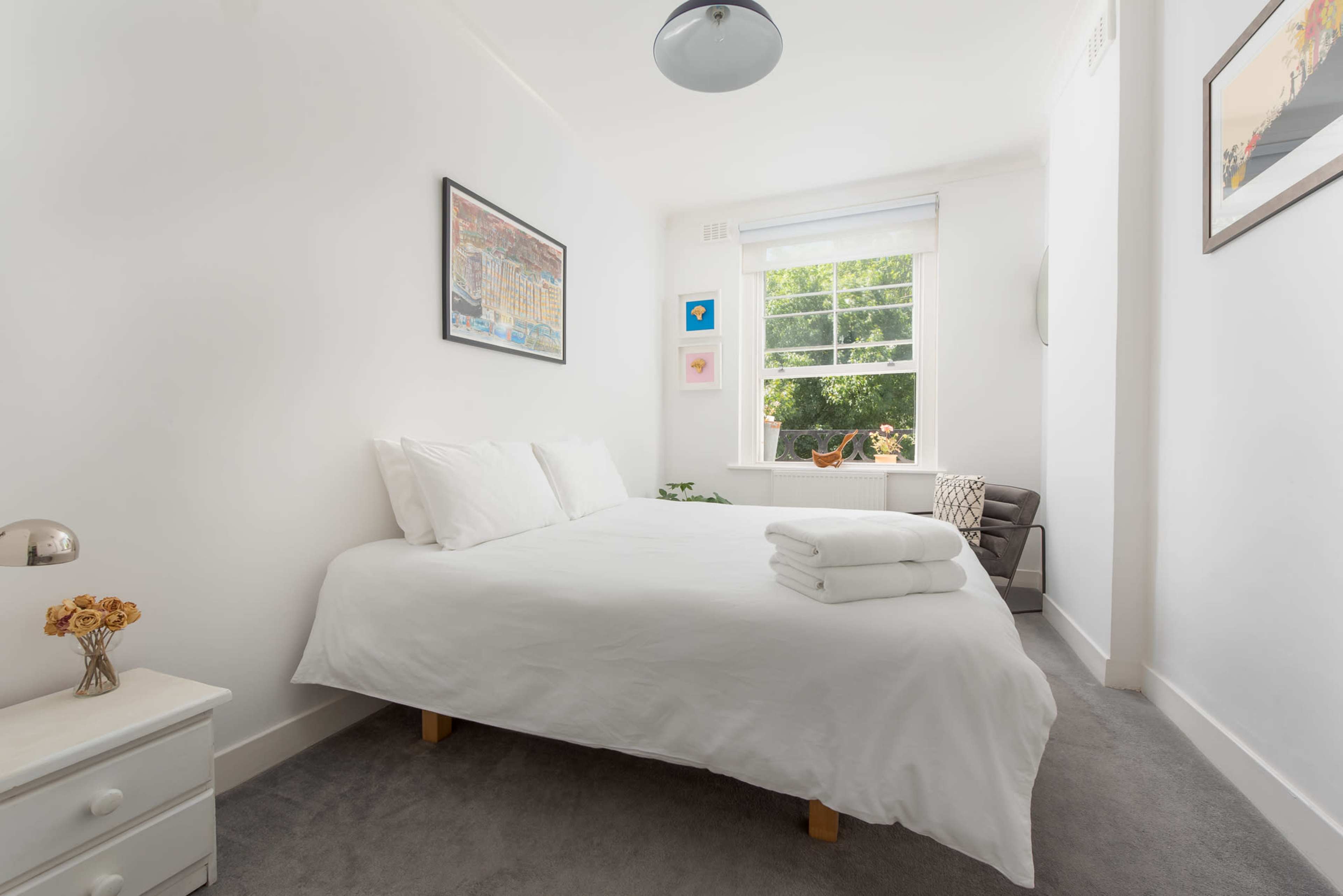 A bright, minimalist bedroom featuring a double bed with white linens, a small side table, and a window with views of greenery.