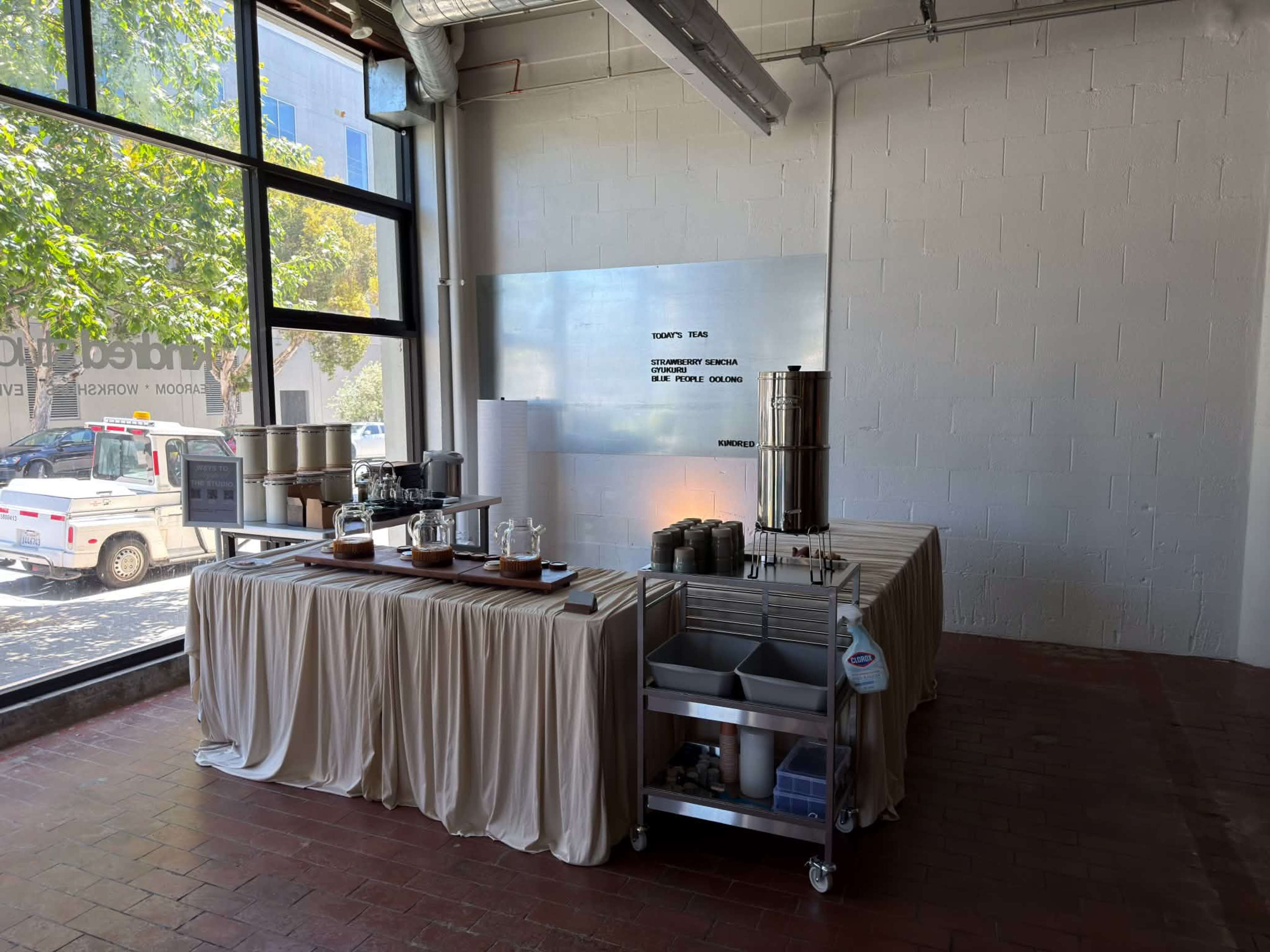 A light-filled room features a neatly arranged table with various containers and utensils on a draped surface, alongside large windows that provide views of greenery outside.