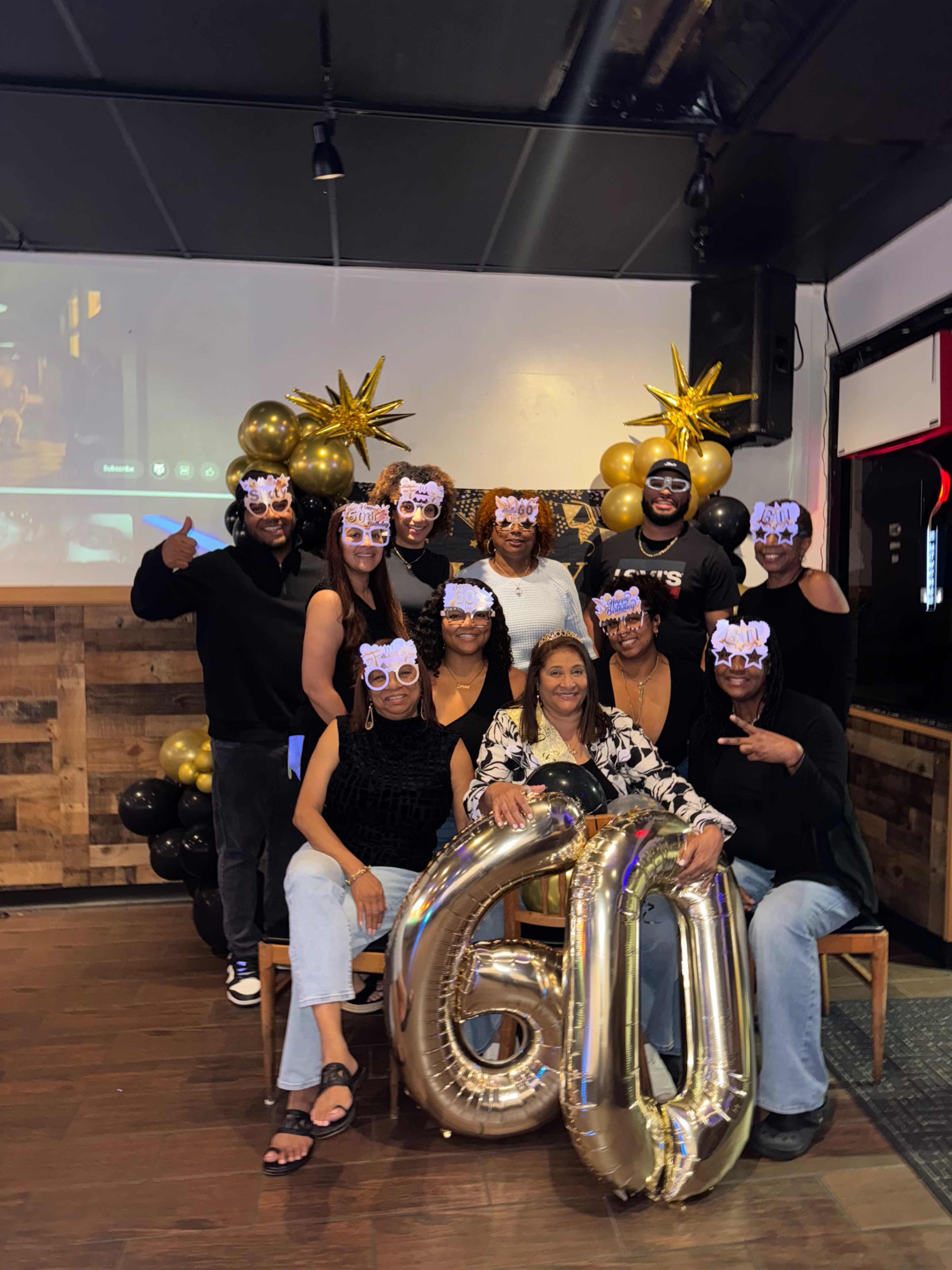 A group of ten people, wearing festive masks, poses together in a decorated venue with gold and black balloons celebrating a 60th birthday.