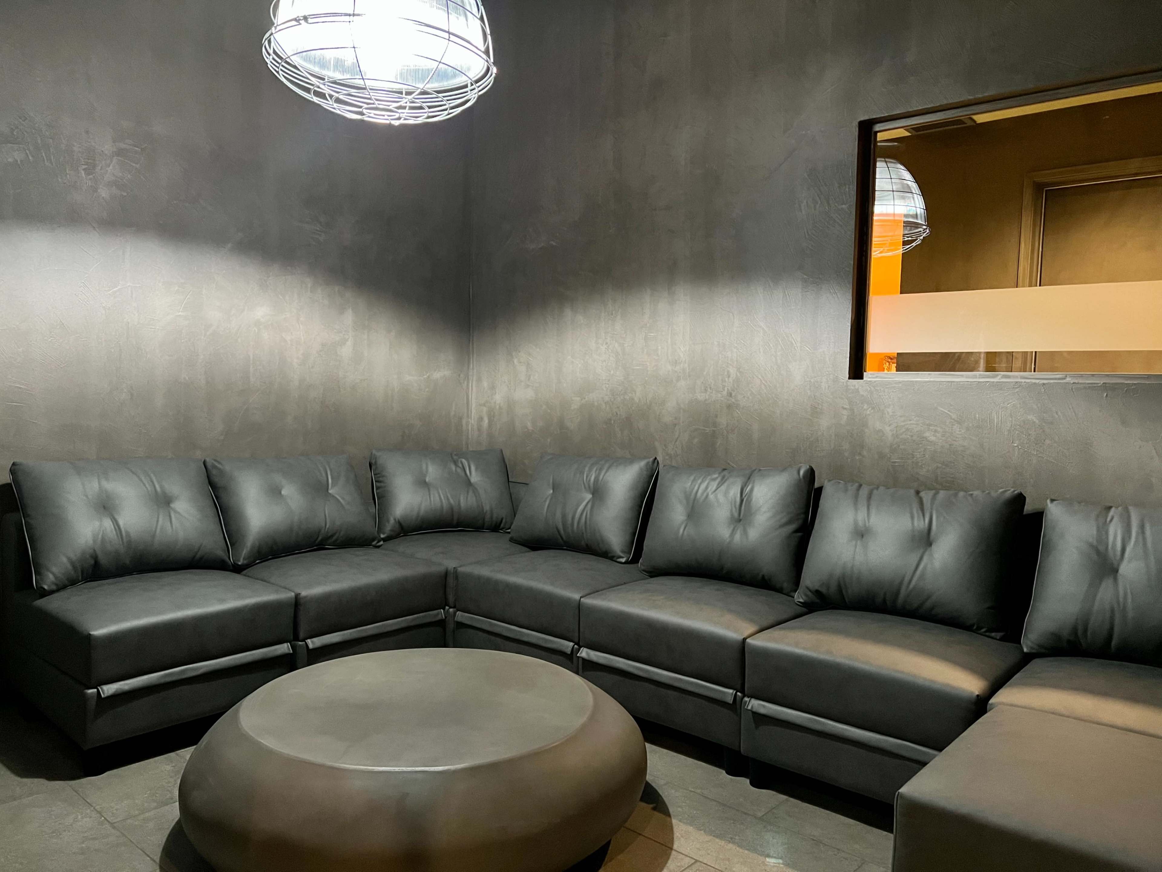 A large, black sectional sofa forms a semi-circle around a round coffee table in a dimly lit room with a wire pendant light overhead.