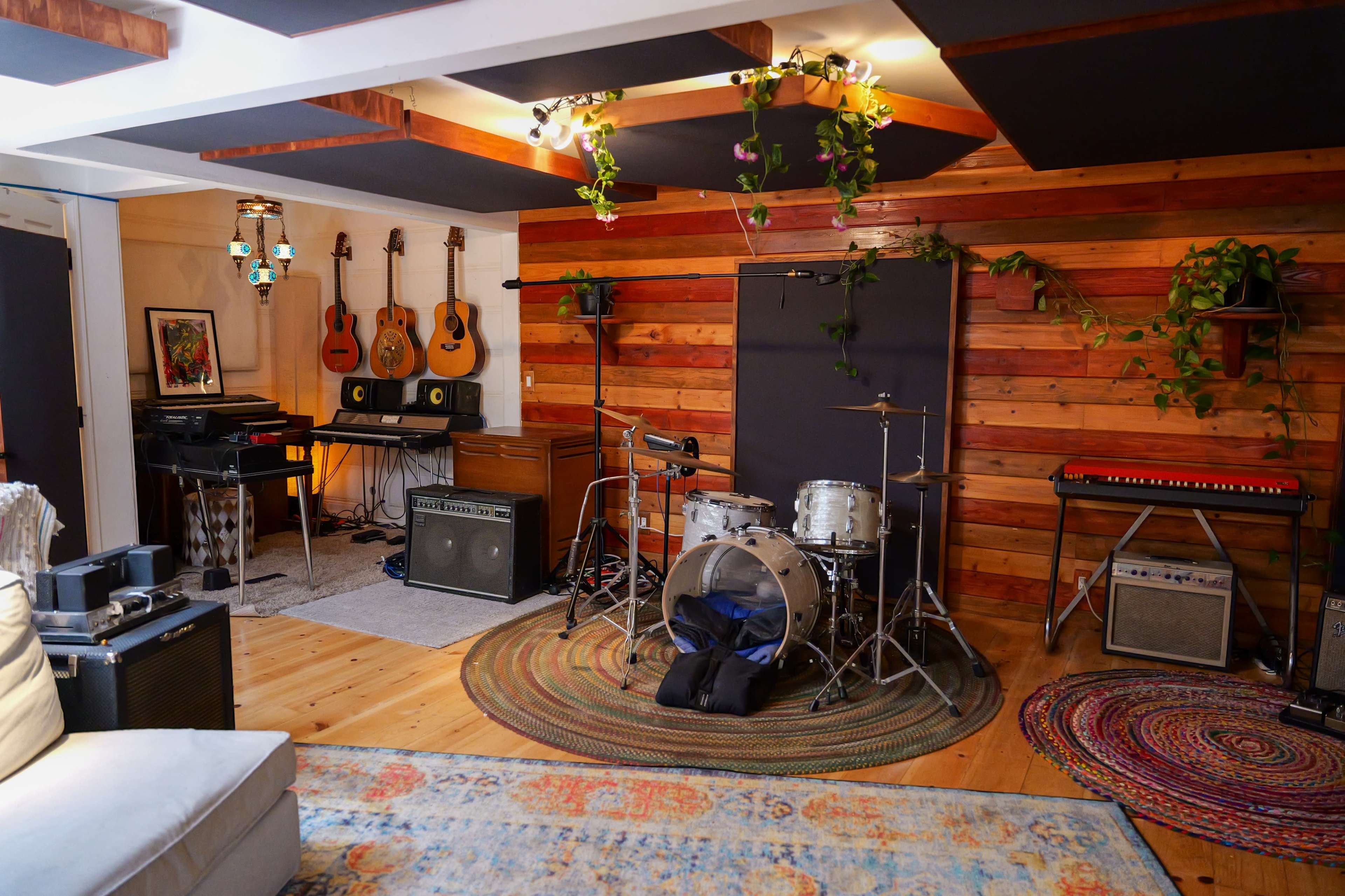 A music practice room featuring drums, guitars, keyboards, amplifiers, and decorative plants against wooden walls.