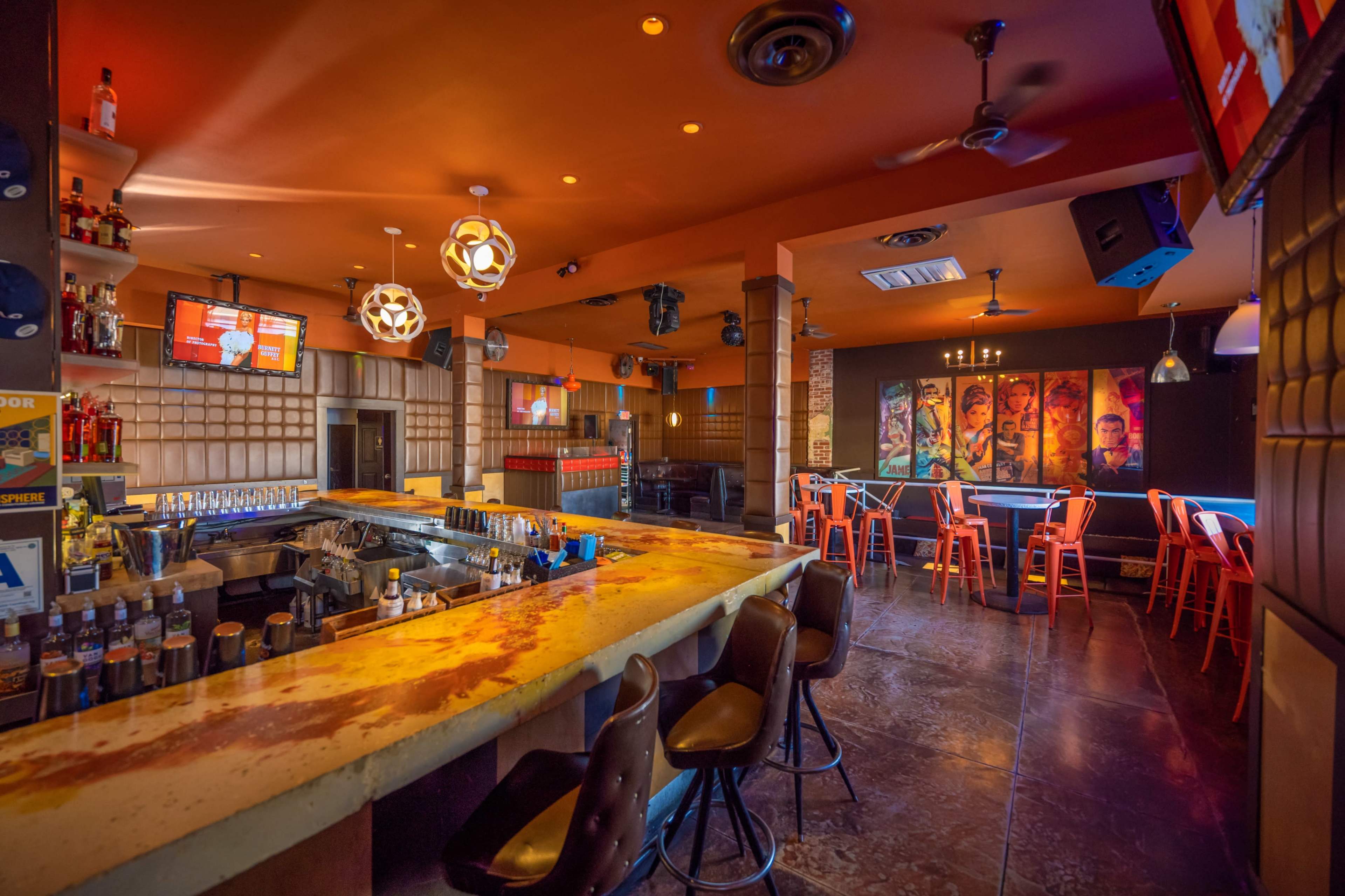 The image shows a stylish bar with a vibrant orange ceiling, a long countertop, and colorful seating arrangements.