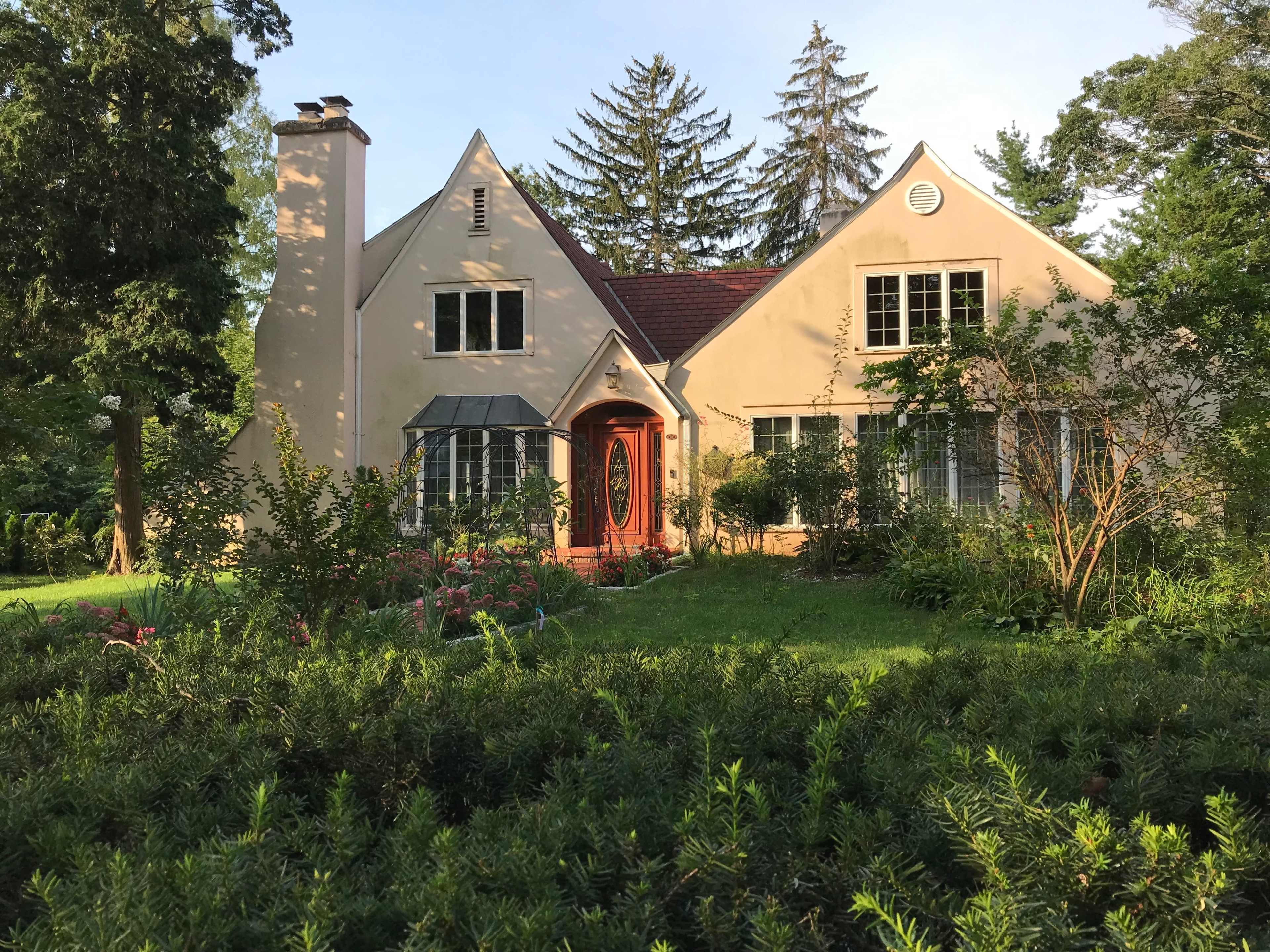 The image shows a two-story house with a sloped roof, surrounded by trees and a garden.