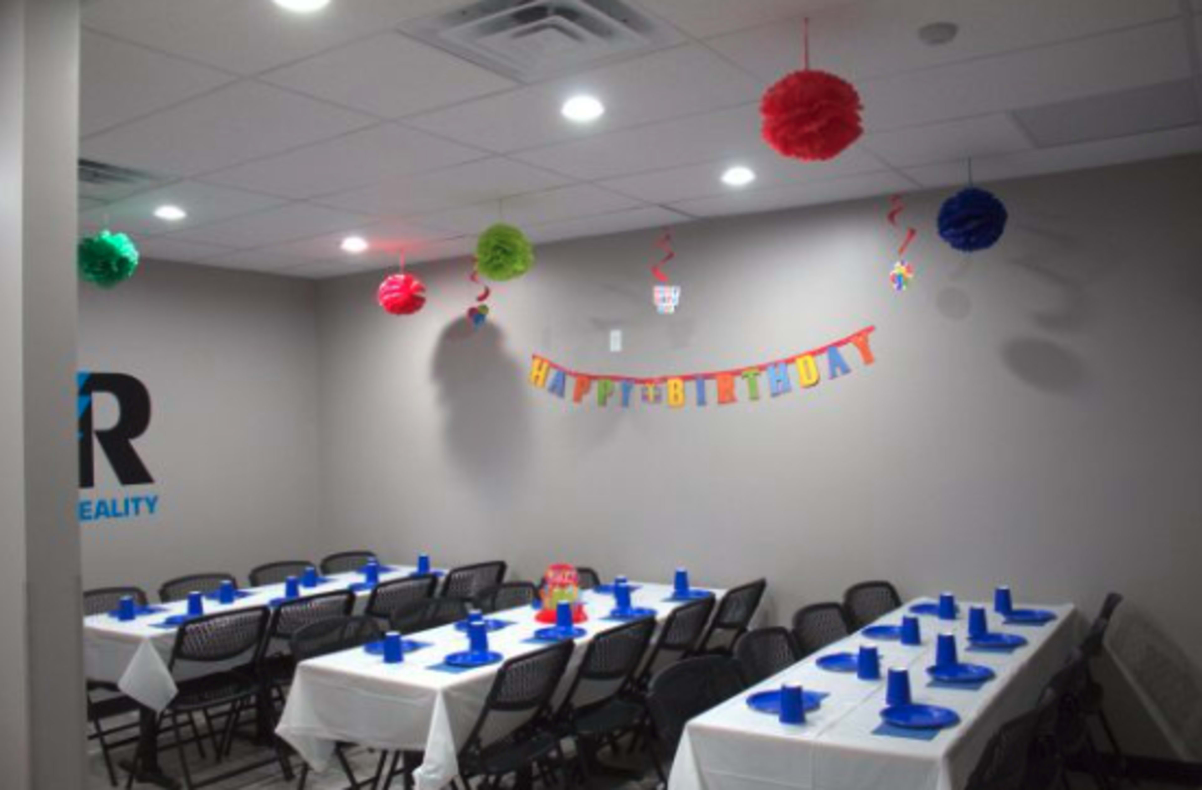 A decorated party room features tables with blue plates and cups, a "Happy Birthday" banner, and colorful hanging decorations.