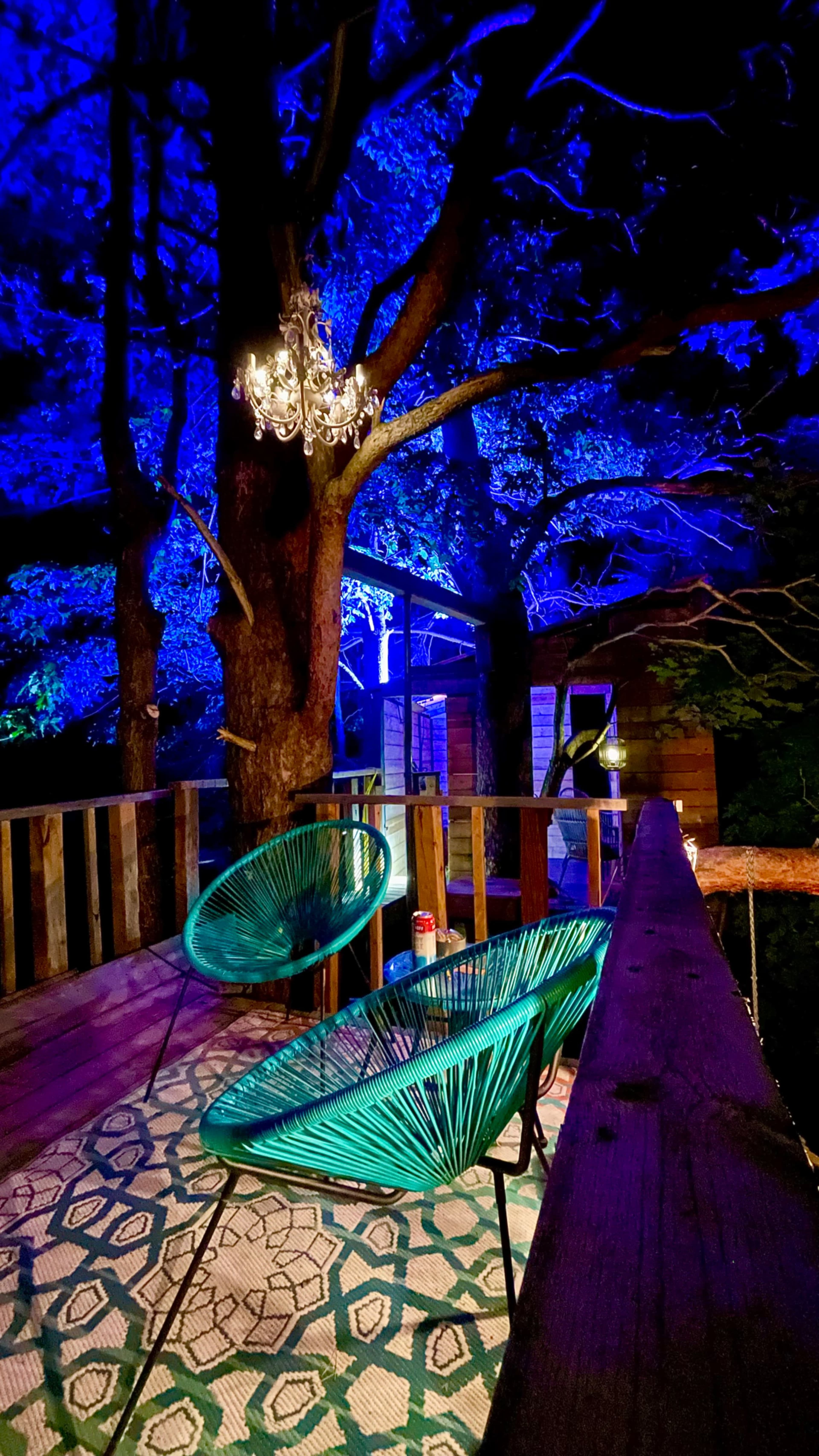 A terrace surrounded by trees features two green chairs and a chandelier, illuminated by blue lighting at night.