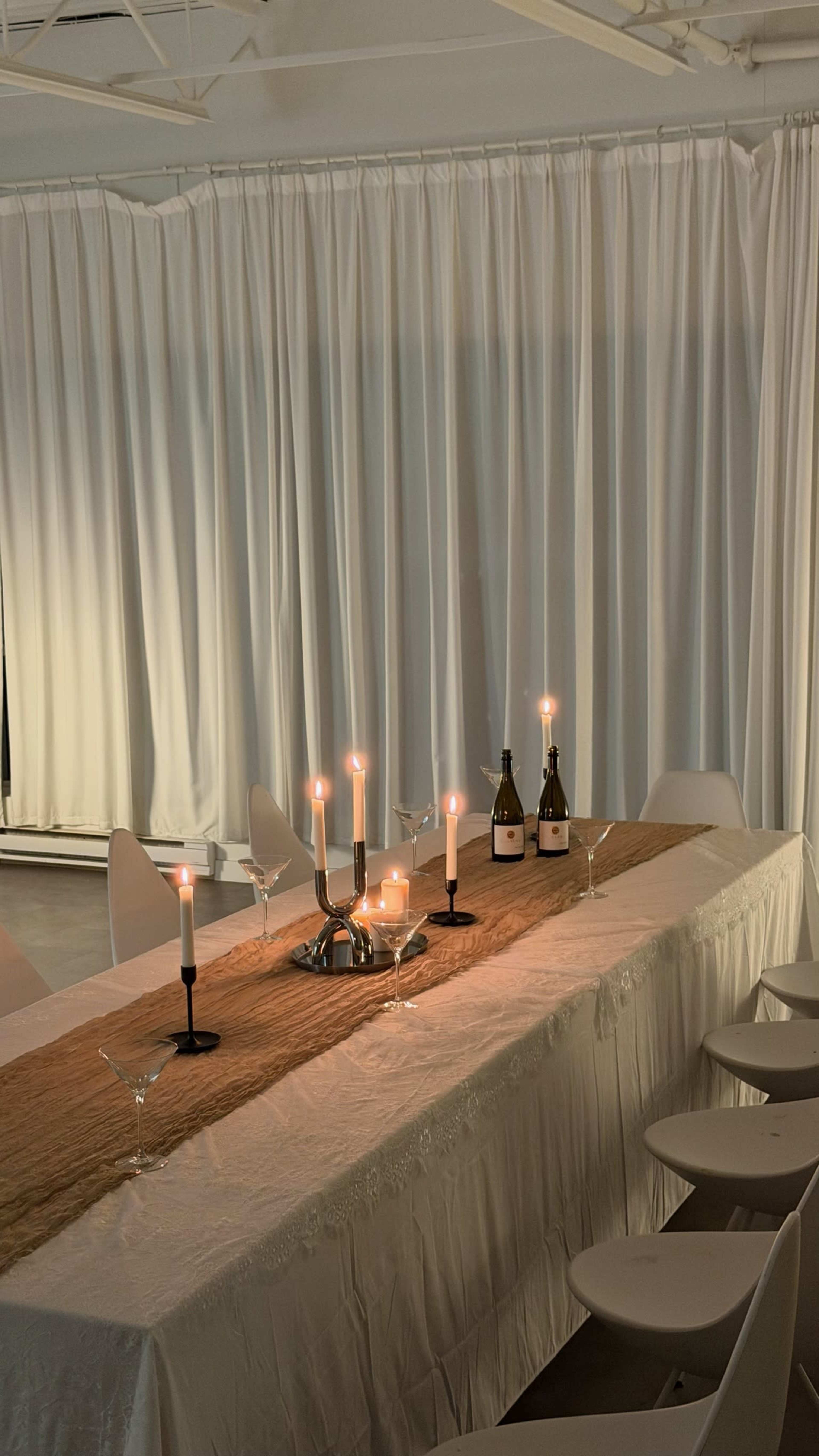 A long dining table is set with candles, a decorative centerpiece, and two bottles in a well-lit, minimalist room.