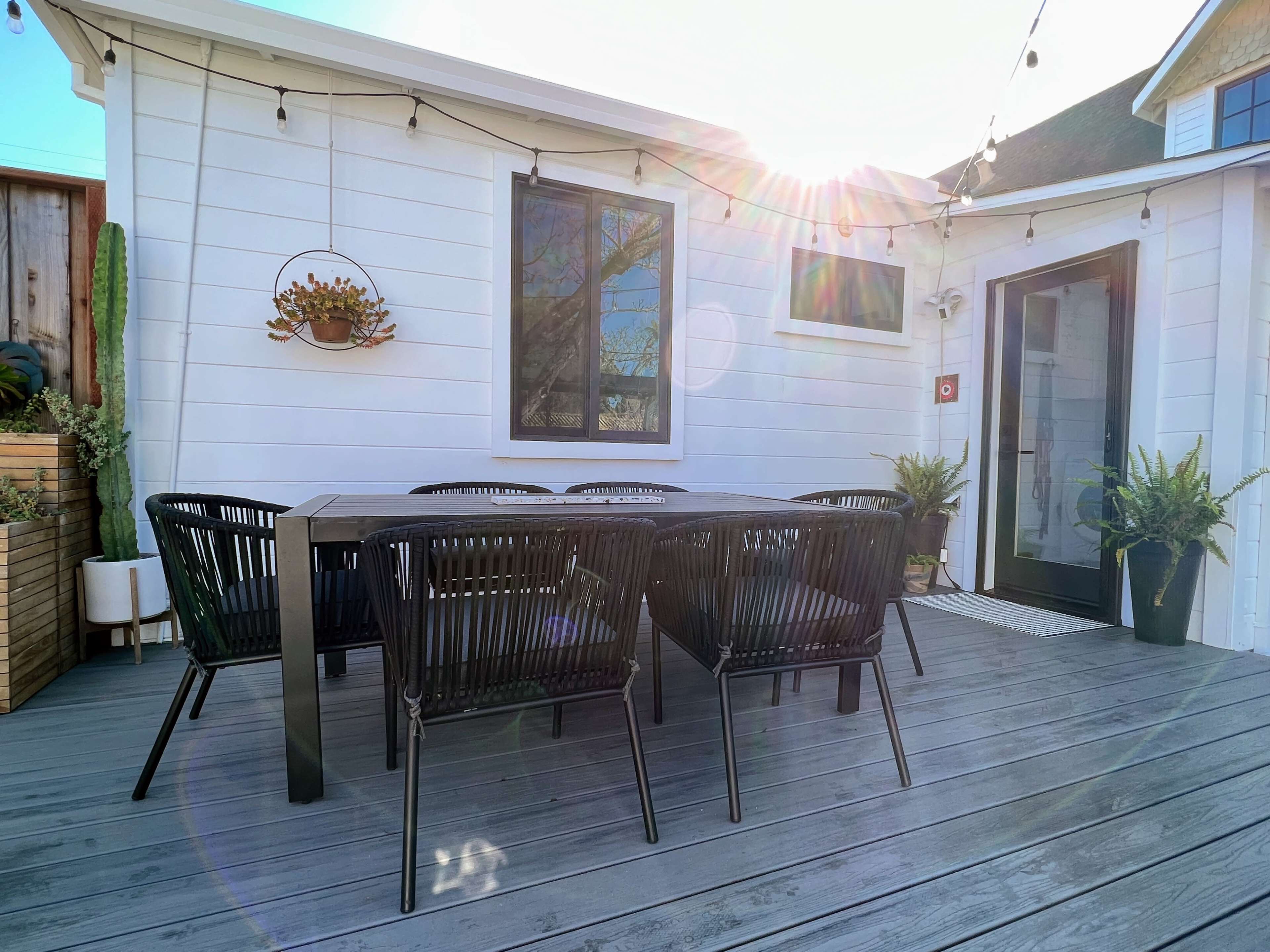 A dining table with black chairs is set up on a wooden deck beside a white house, with sunlight streaming in from the left.