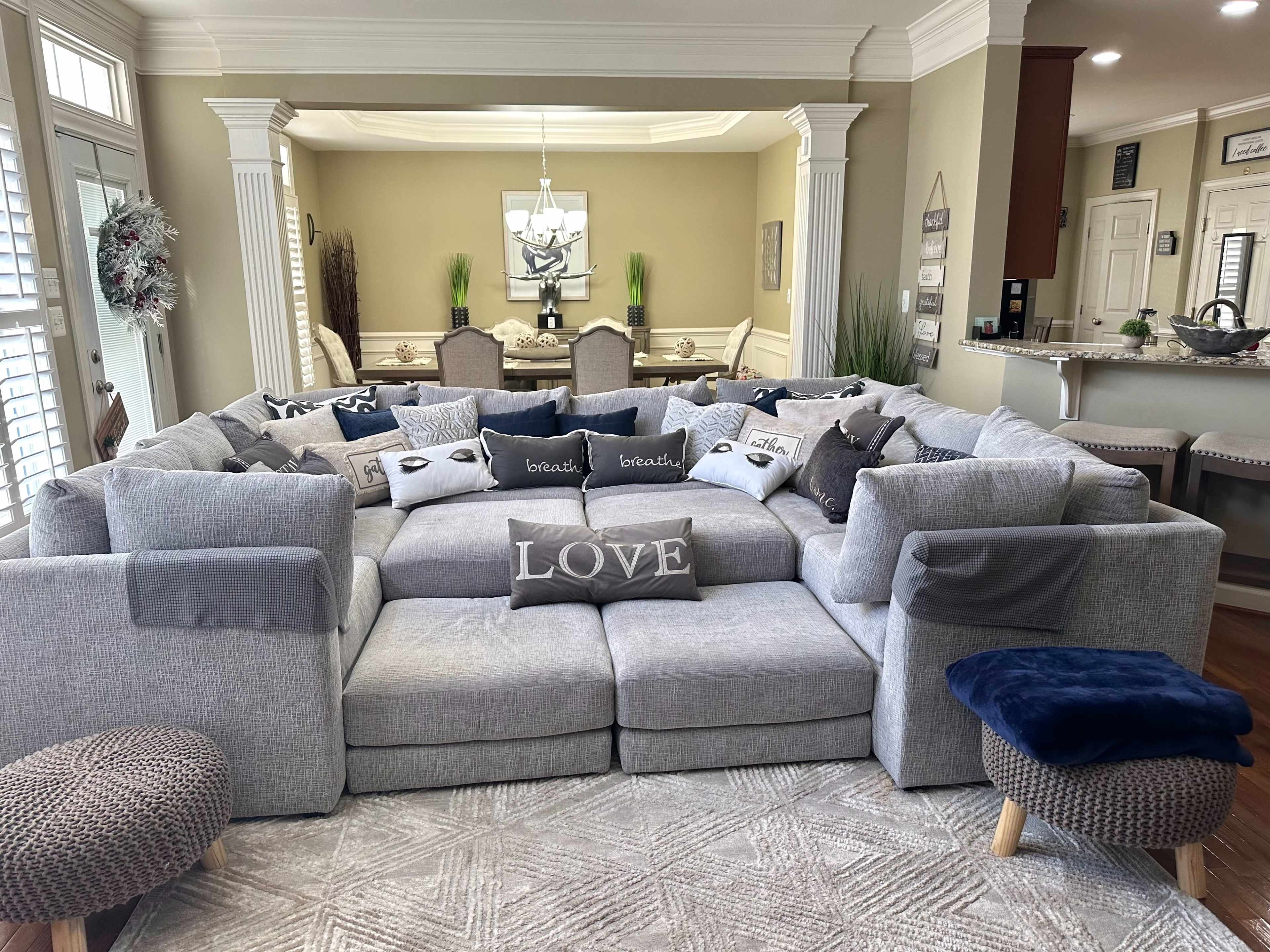 A large, L-shaped sectional sofa is arranged in a cozy living area, surrounded by numerous decorative pillows with various designs, including the word "LOVE."