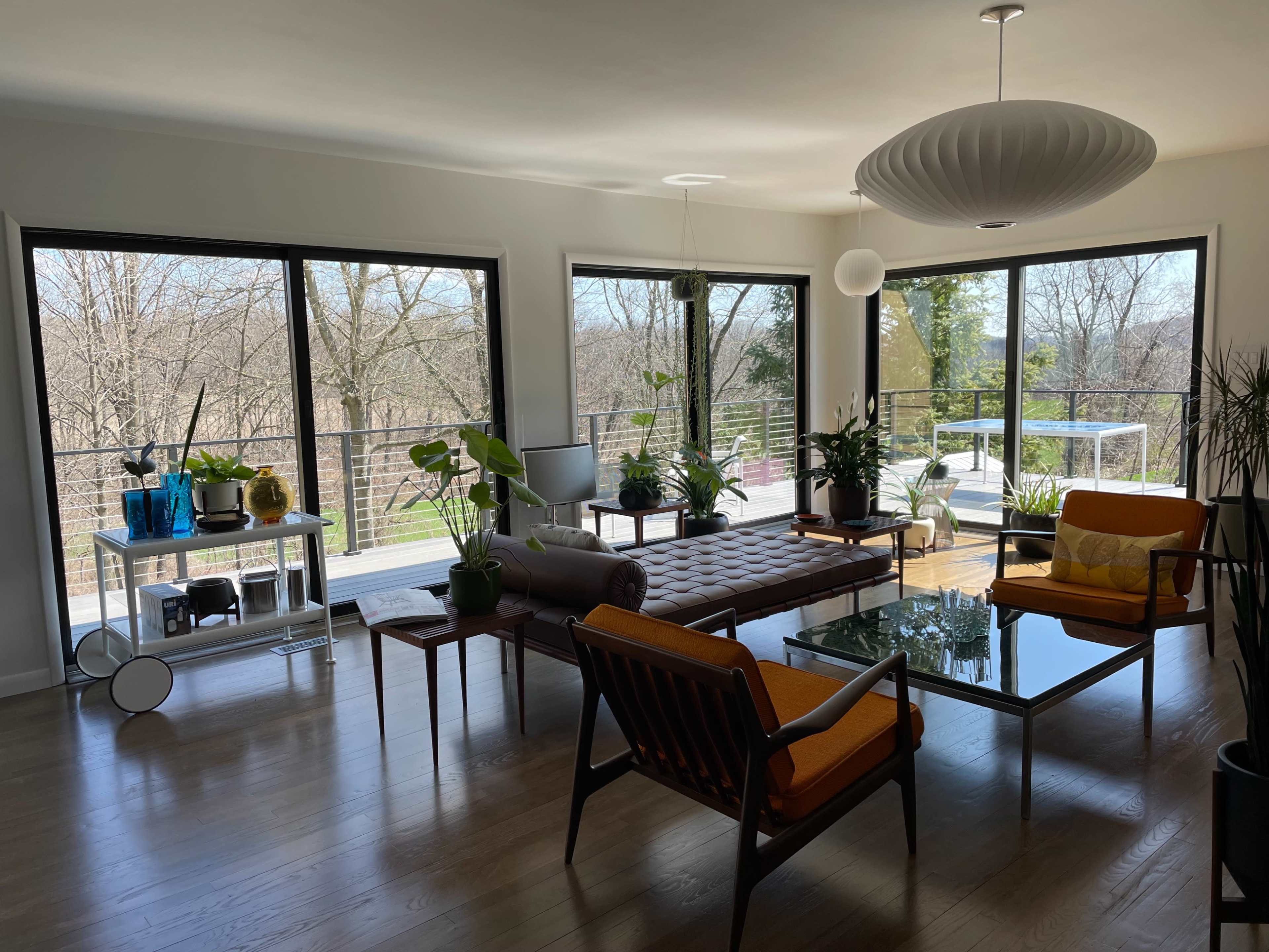 A modern living room with large windows overlooking a wooded area, furnished with a brown sofa, orange chairs, and various potted plants.