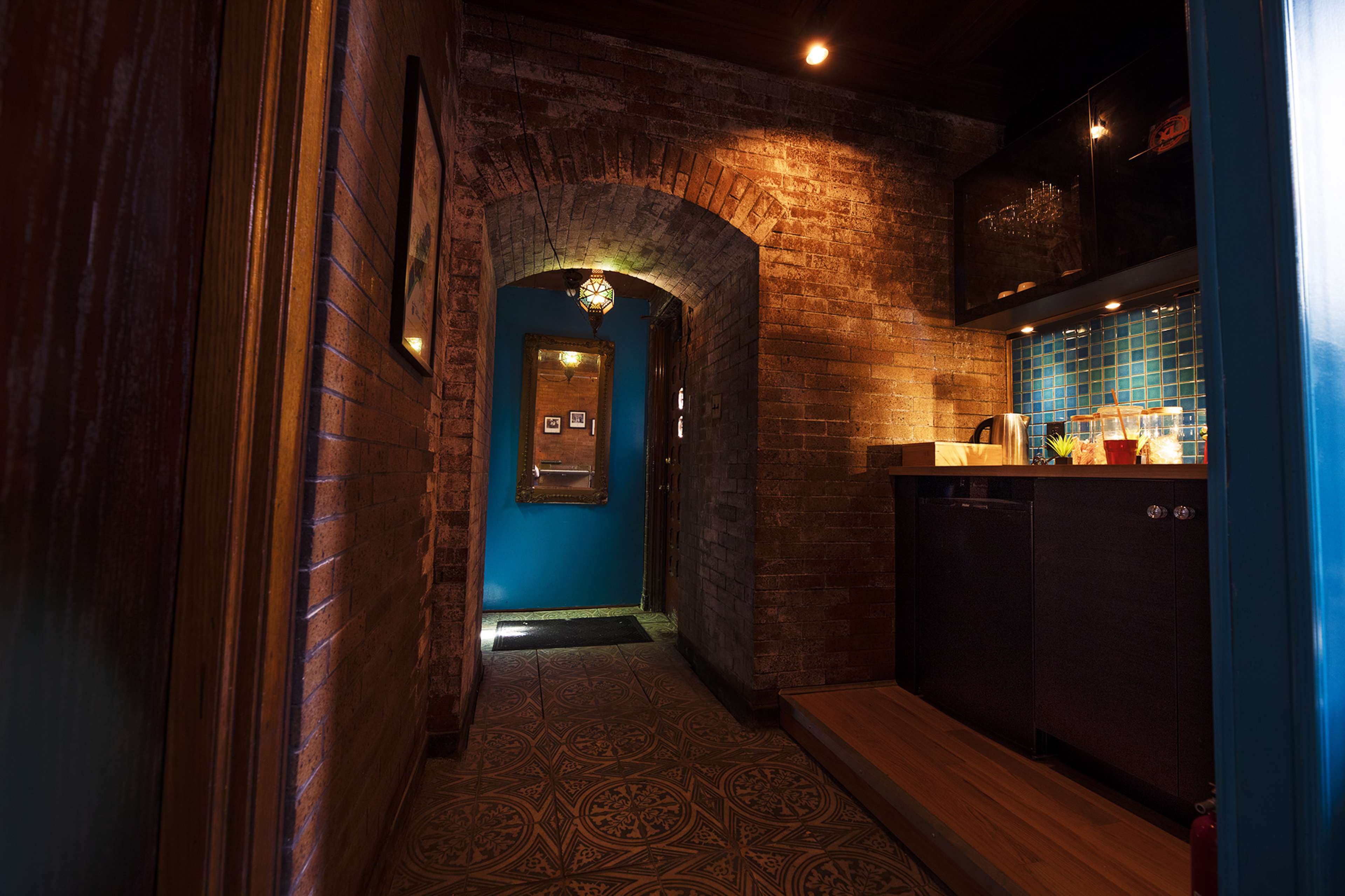 A narrow hallway with exposed brick walls leading to a blue door, featuring a kitchen area with a countertop and mosaic backsplash.