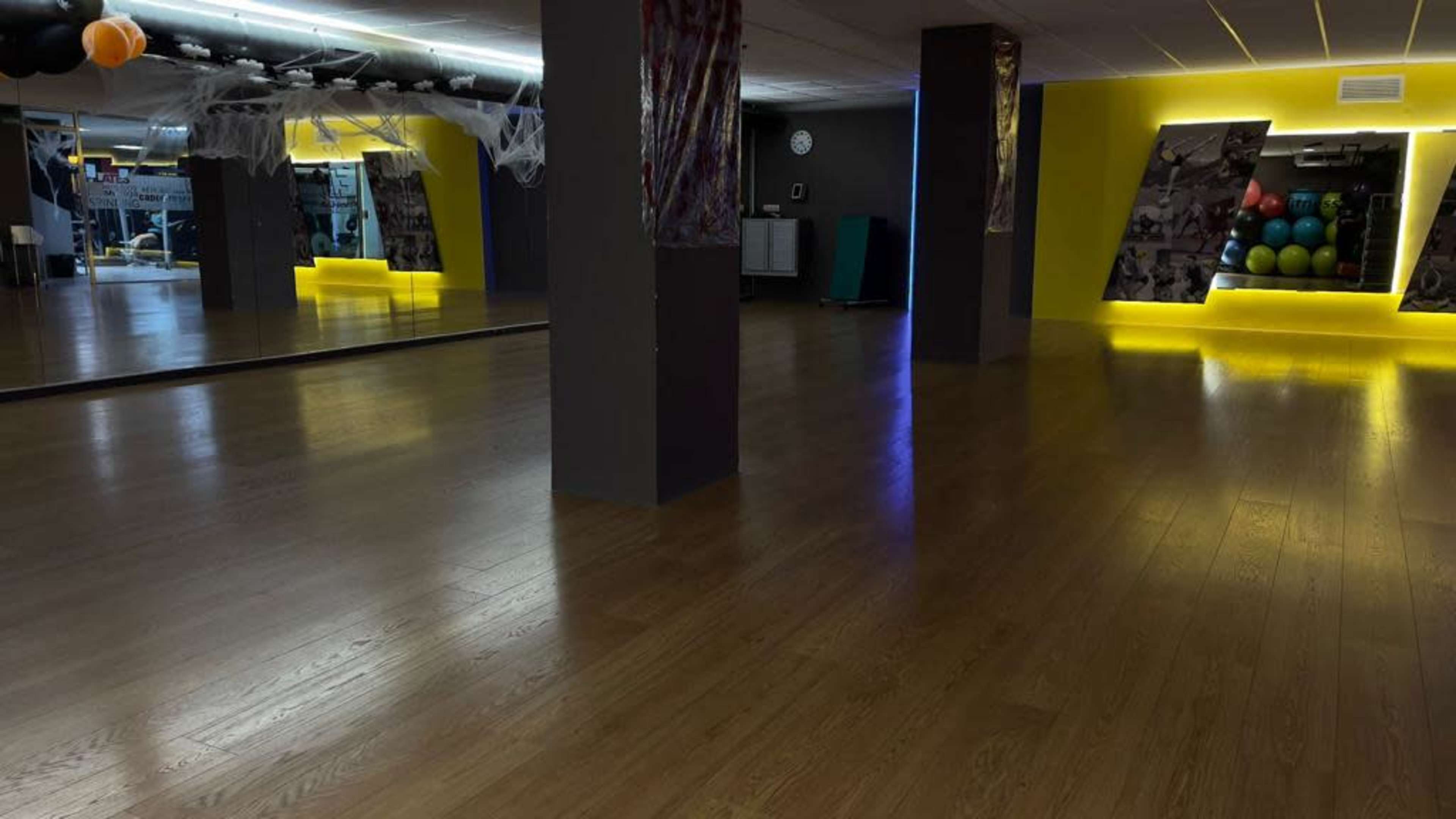 The image shows an empty fitness studio with wooden flooring, mirrors, and colorful exercise equipment against a brightly lit wall.