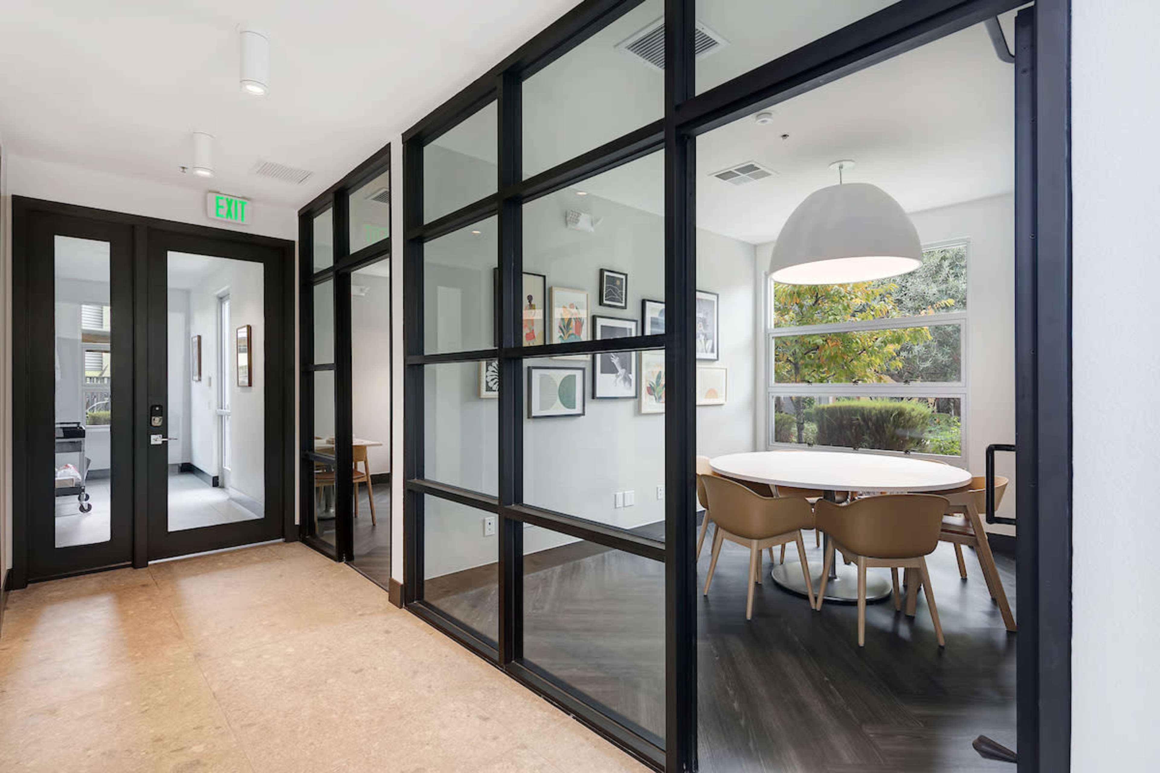 A hallway features black-framed glass walls that lead to a modern meeting room with a round table and chairs.