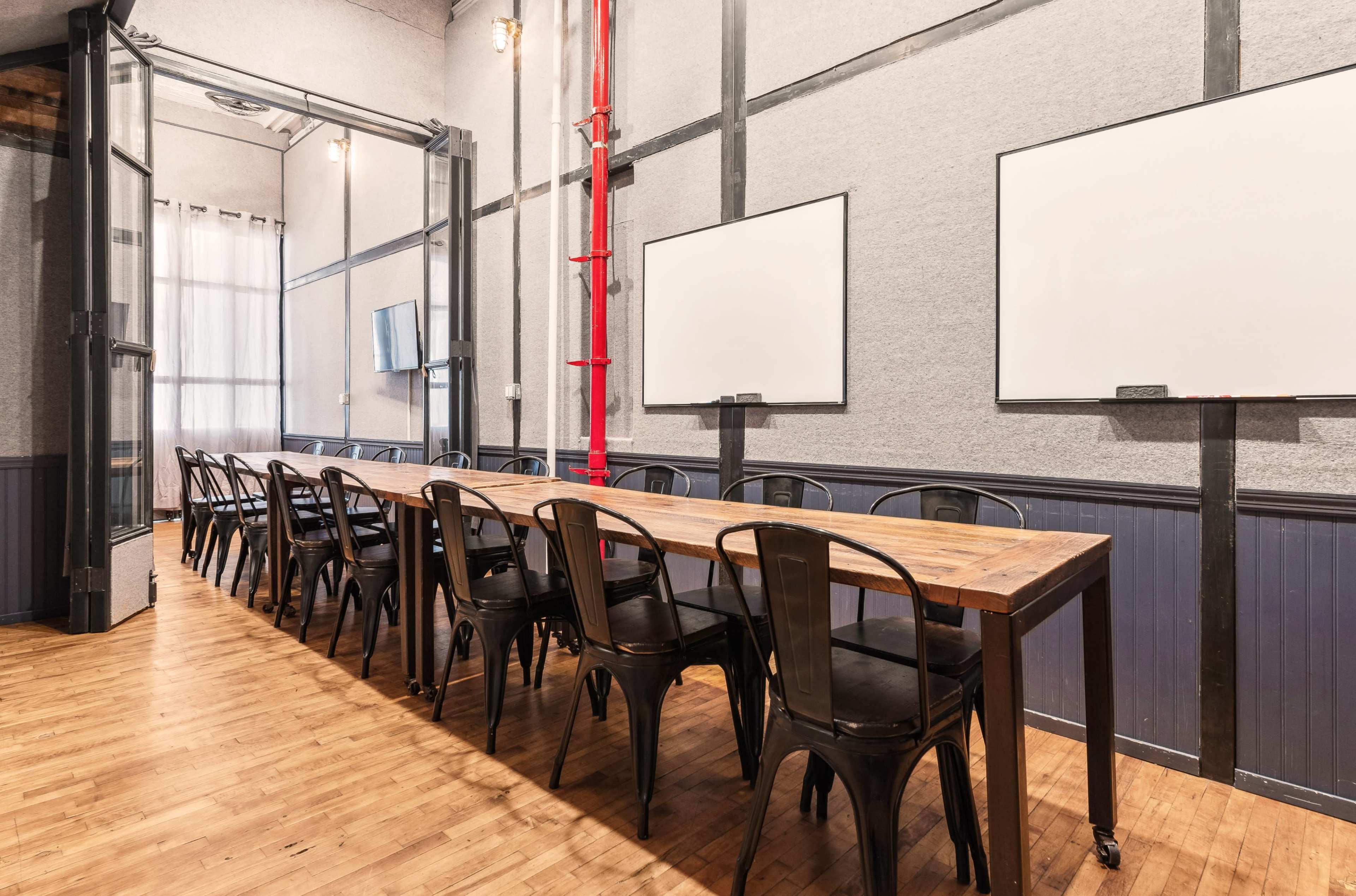 A long wooden table with black chairs is arranged in a meeting room featuring two whiteboards and a large window.