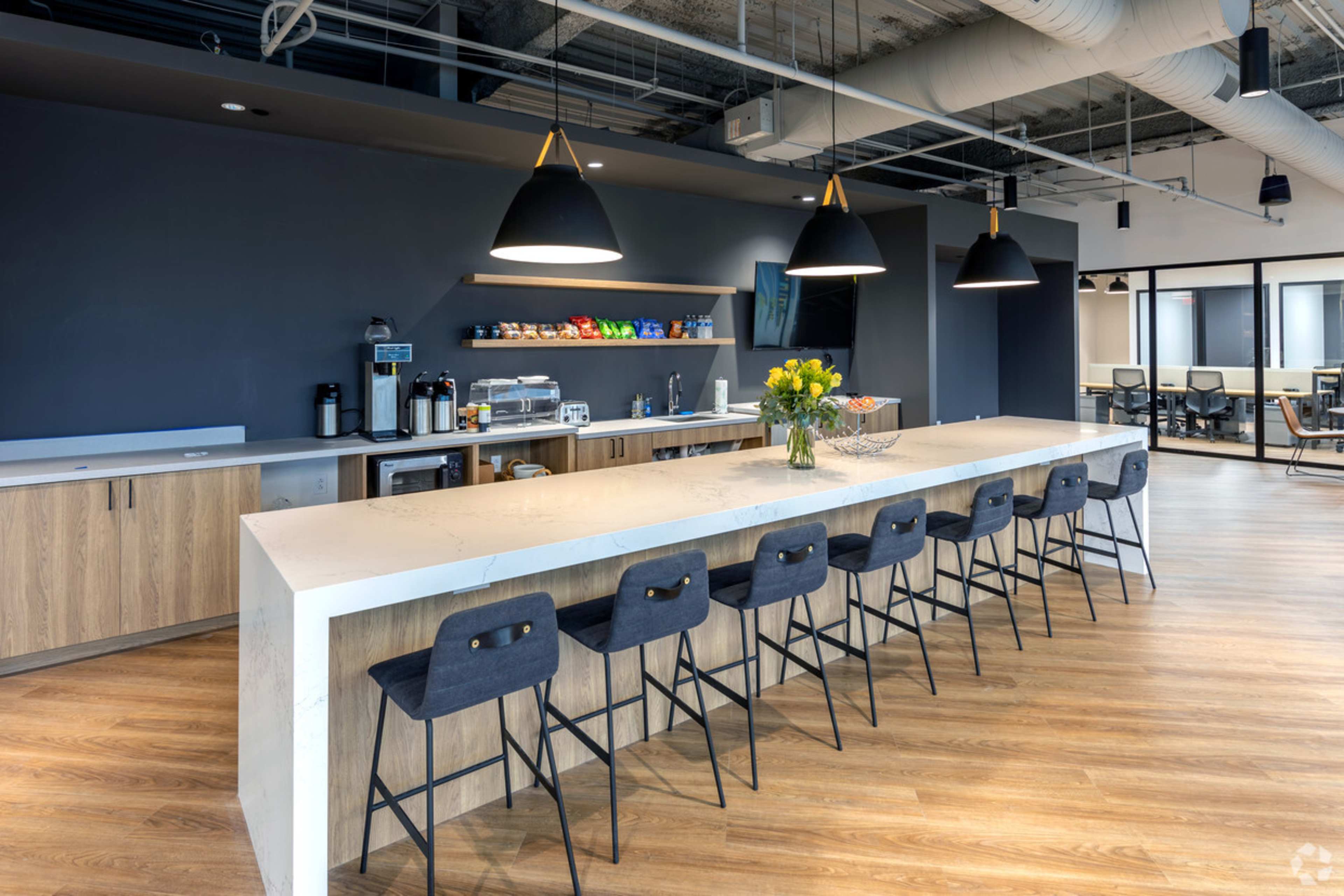 A modern office kitchen area featuring a long island countertop with bar stools and a selection of snacks and beverages in the background.