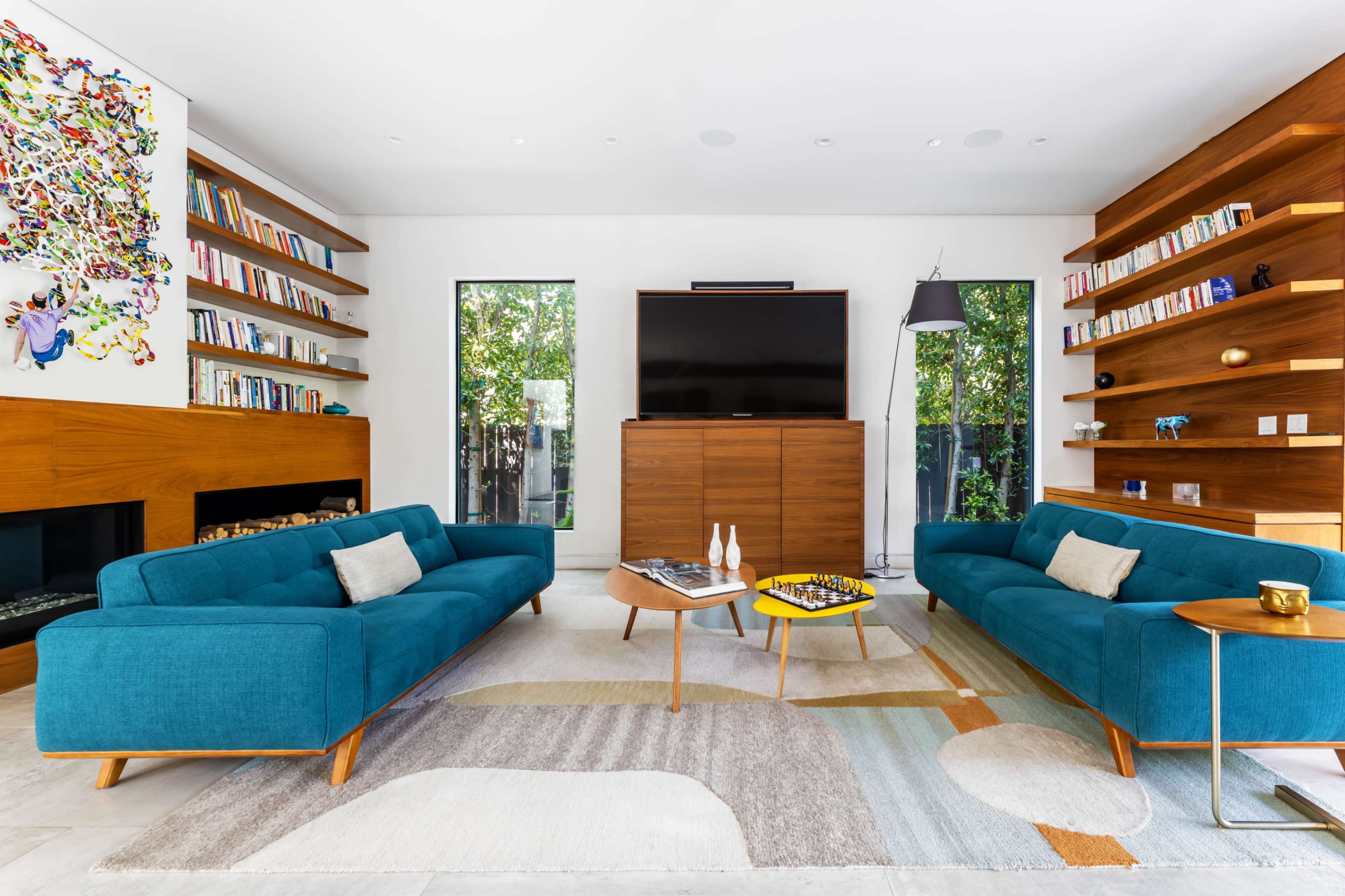 The image shows a modern living room with two teal sofas, a wooden entertainment unit, shelves filled with books, and a large abstract artwork on the wall.