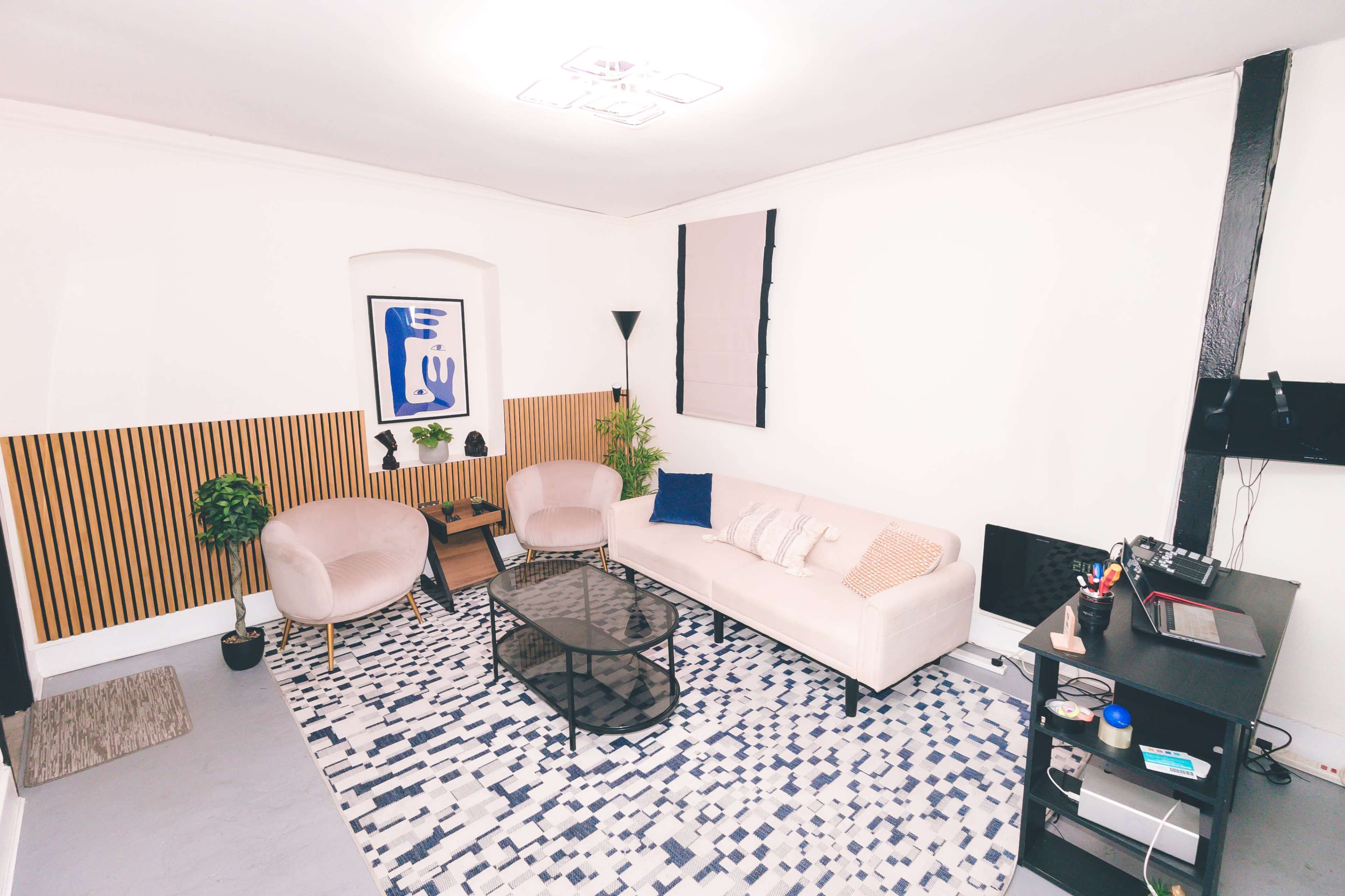 The image shows a modern living room featuring a beige sofa, two armchairs, a coffee table, and a patterned rug, with greenery and simple decor elements against a light-colored wall.