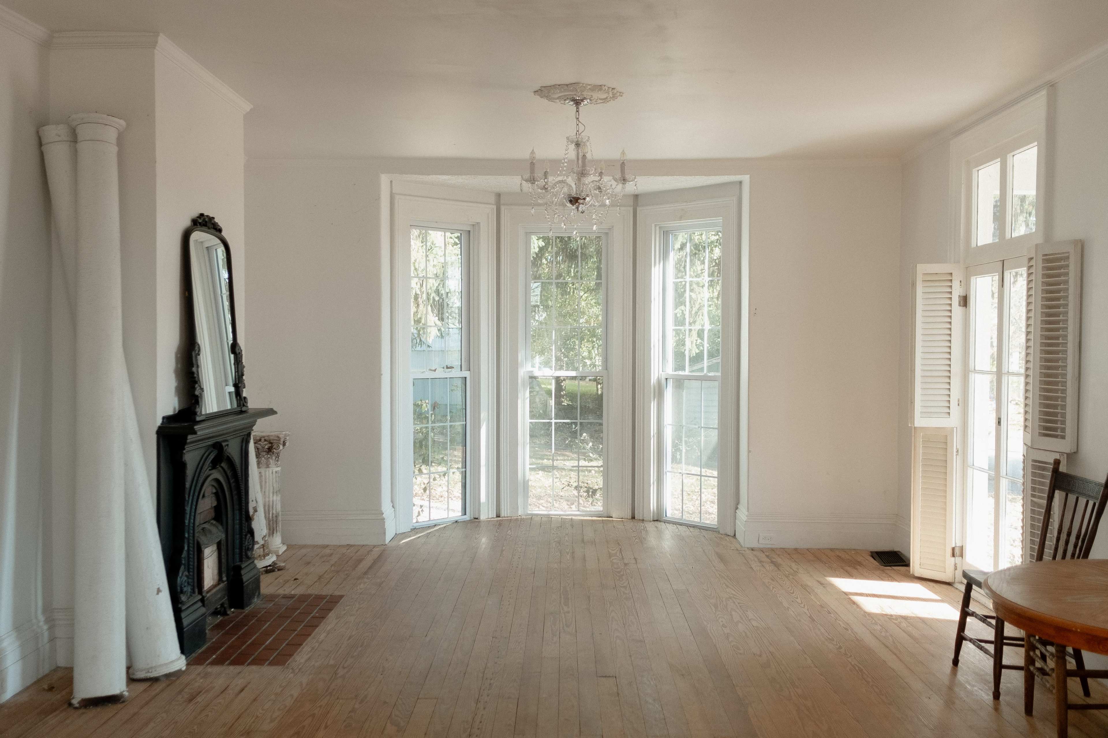 A bright, empty room features large windows, wooden floors, a chandelier, and a decorative fireplace.