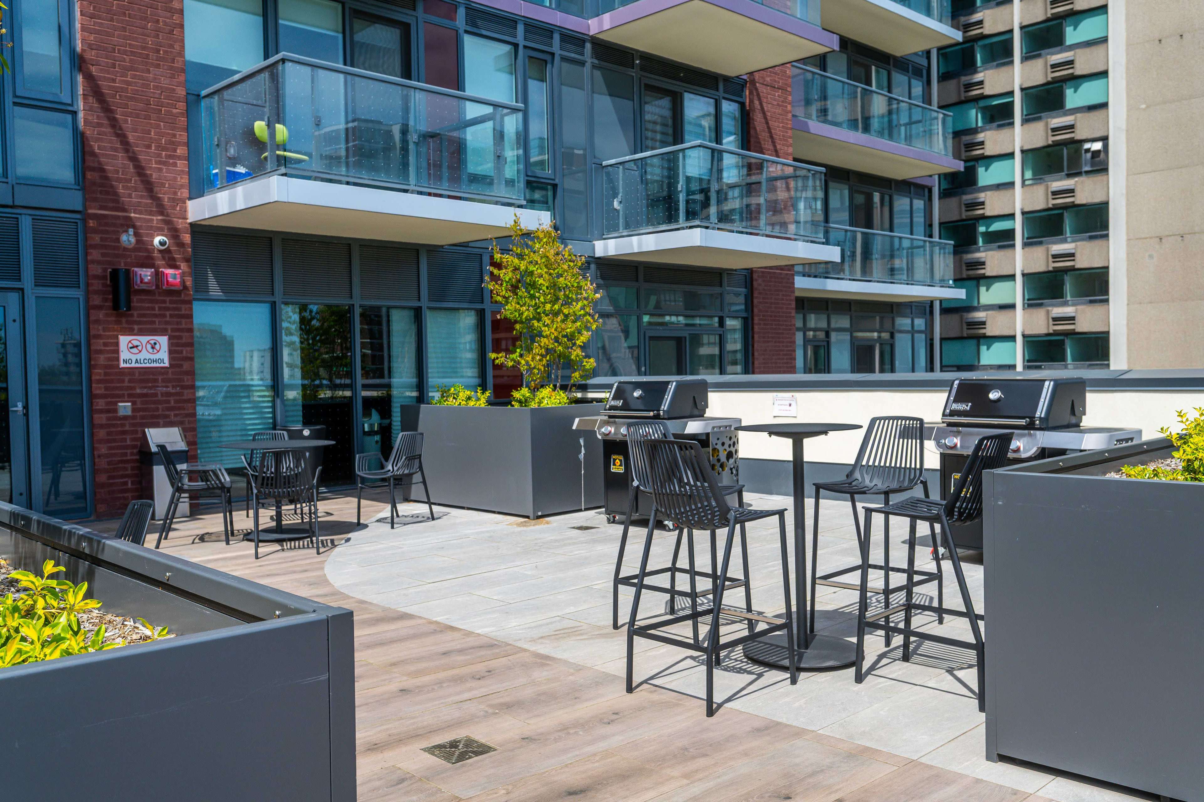 The image shows an outdoor terrace area featuring stainless steel grills, tall tables with stools, and planters in a modern apartment building setting.