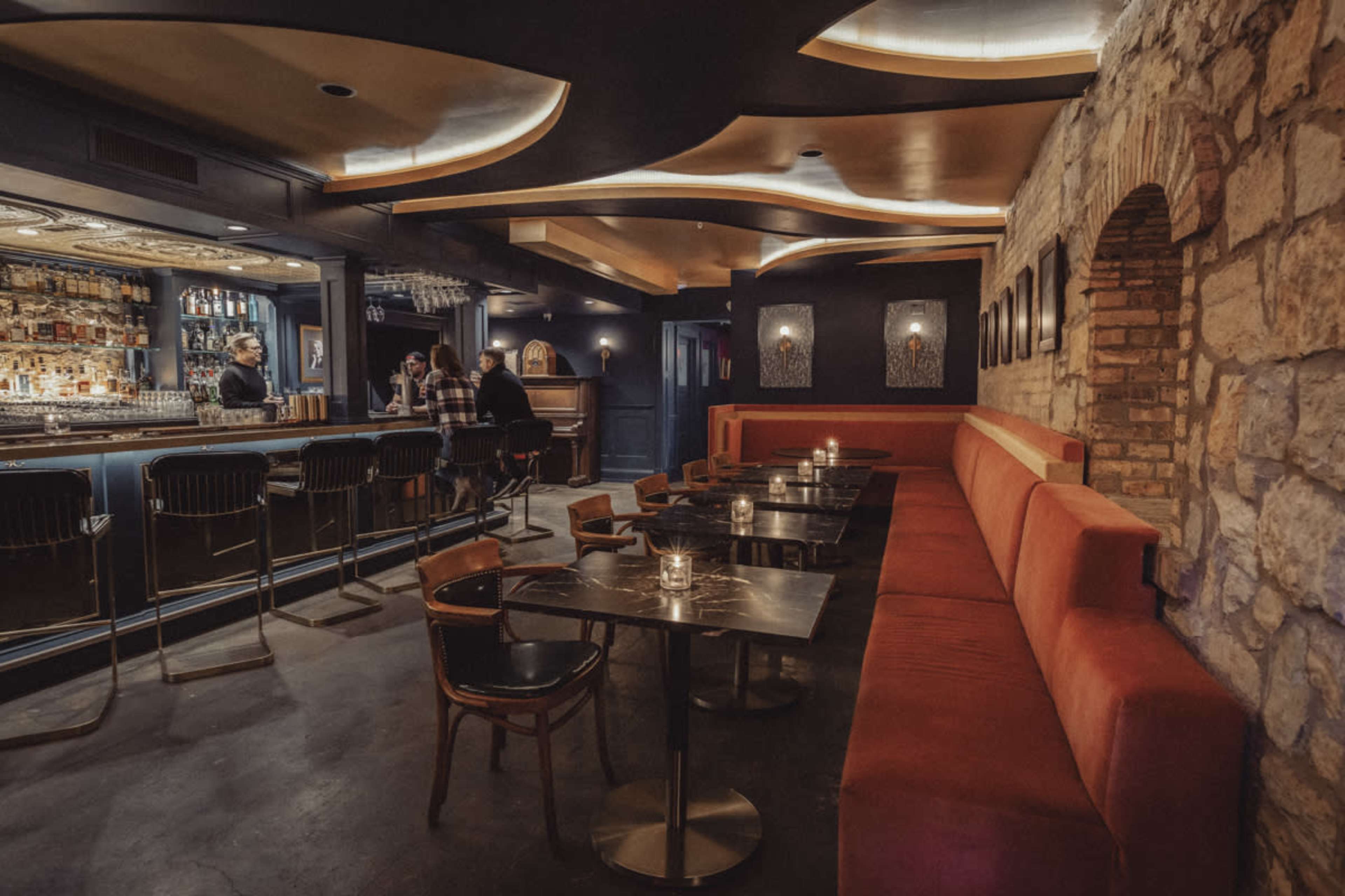 A dimly lit bar interior featuring a curved ceiling, a stone wall, and a seating area with red velvet booths and small tables.