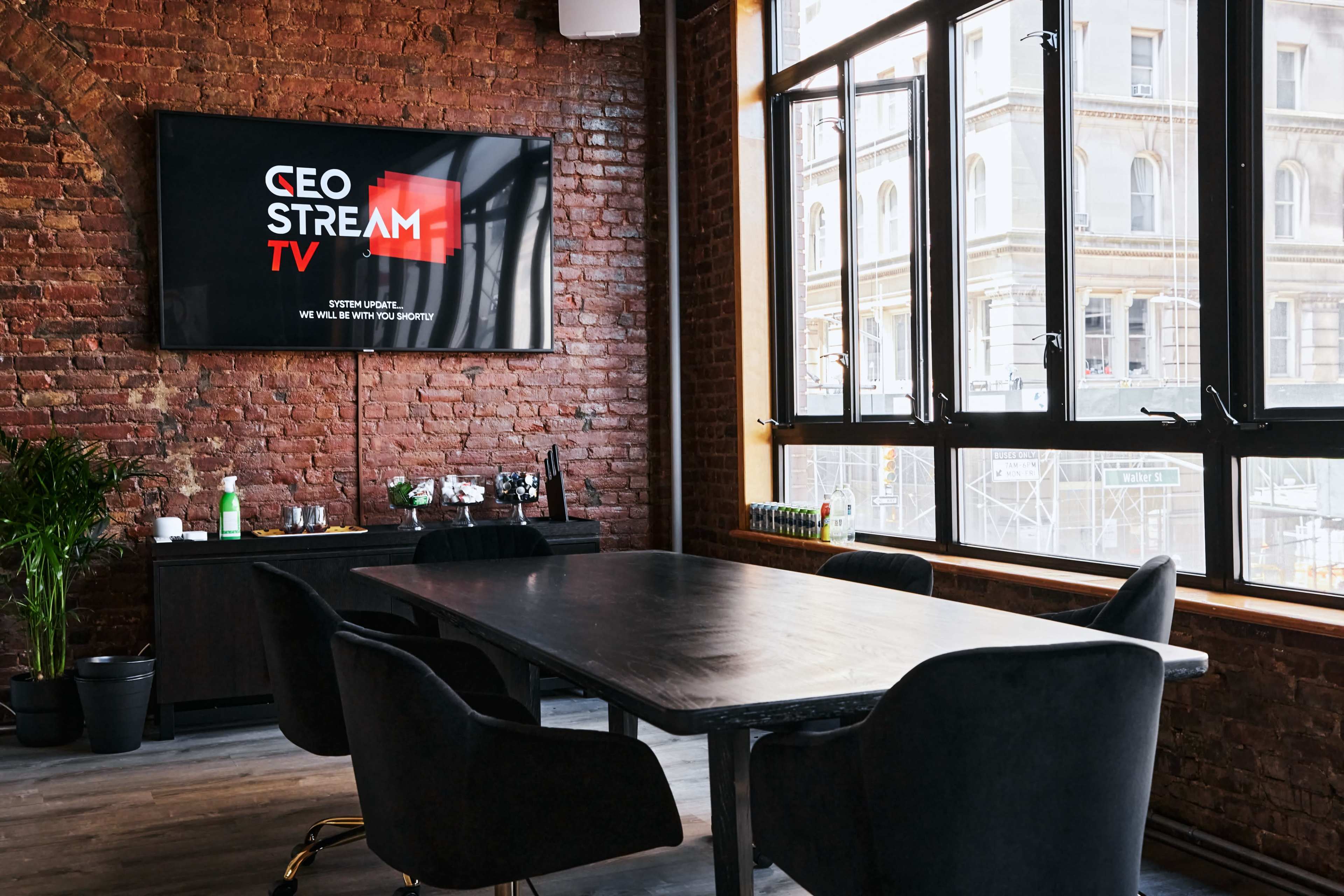 A sleek conference room features a large black table surrounded by chairs, with a television displaying the logo for "CEO Stream TV" on a brick wall.