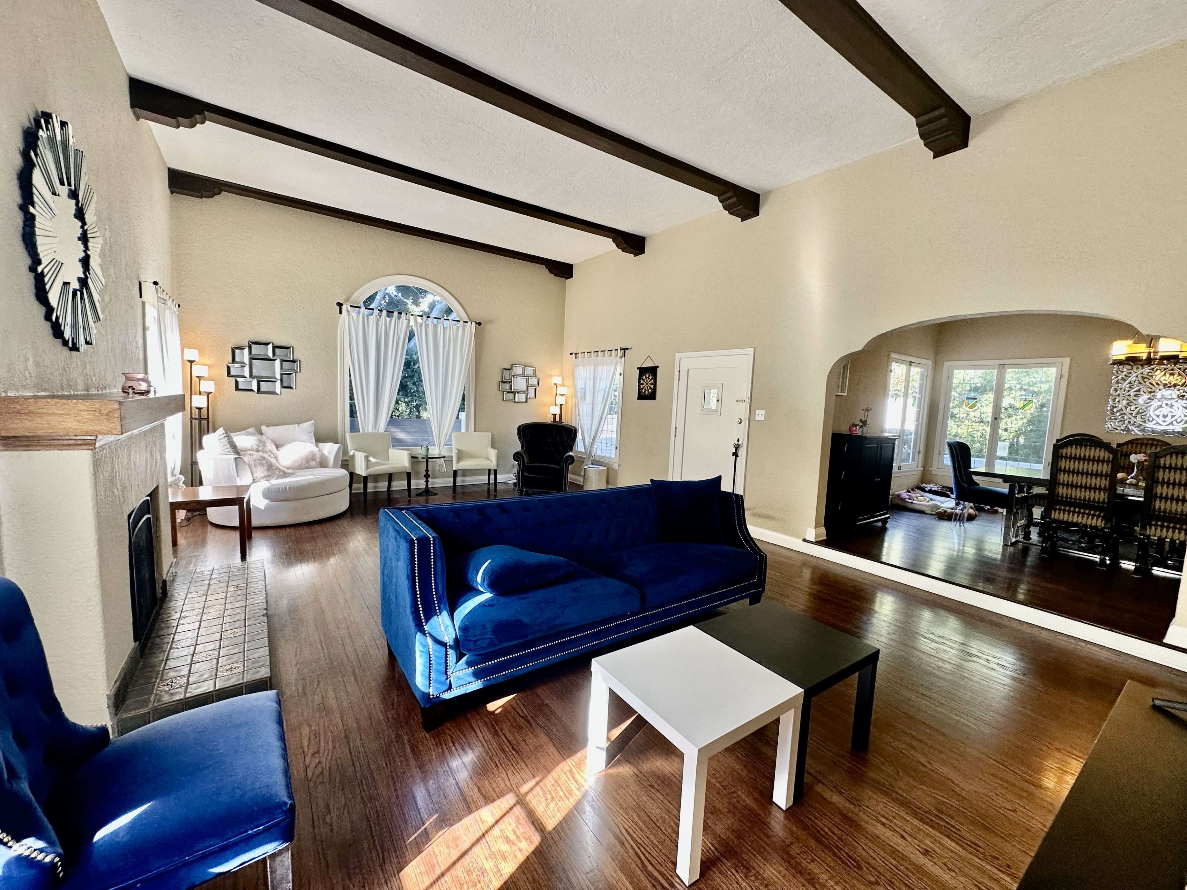 The image shows a spacious living room with a blue sofa, a white coffee table, and multiple seating areas, featuring large windows and warm wood flooring.