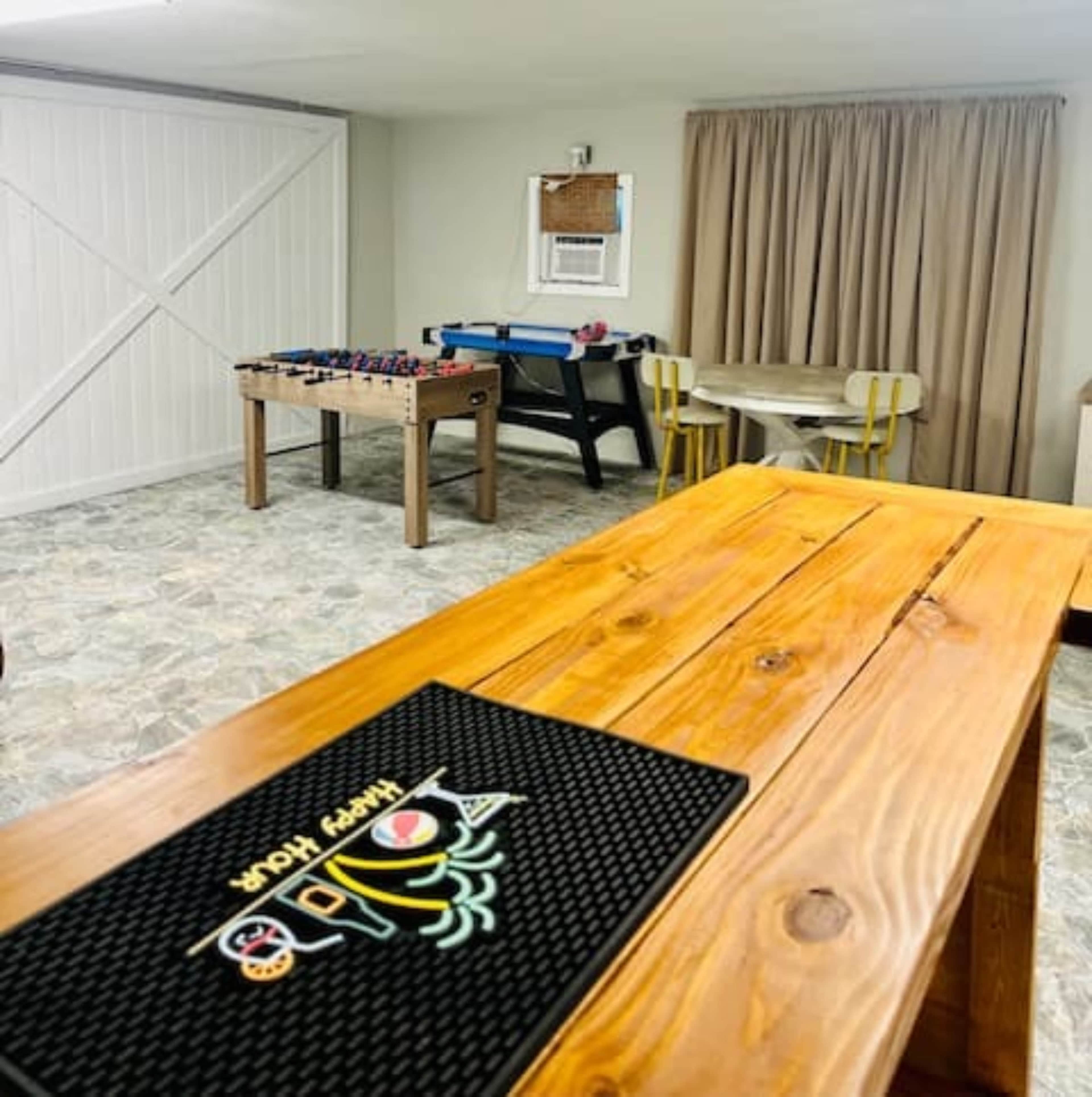 The image shows a game room with a foosball table, a pool table, a round dining table with yellow chairs, and a wooden bar counter.