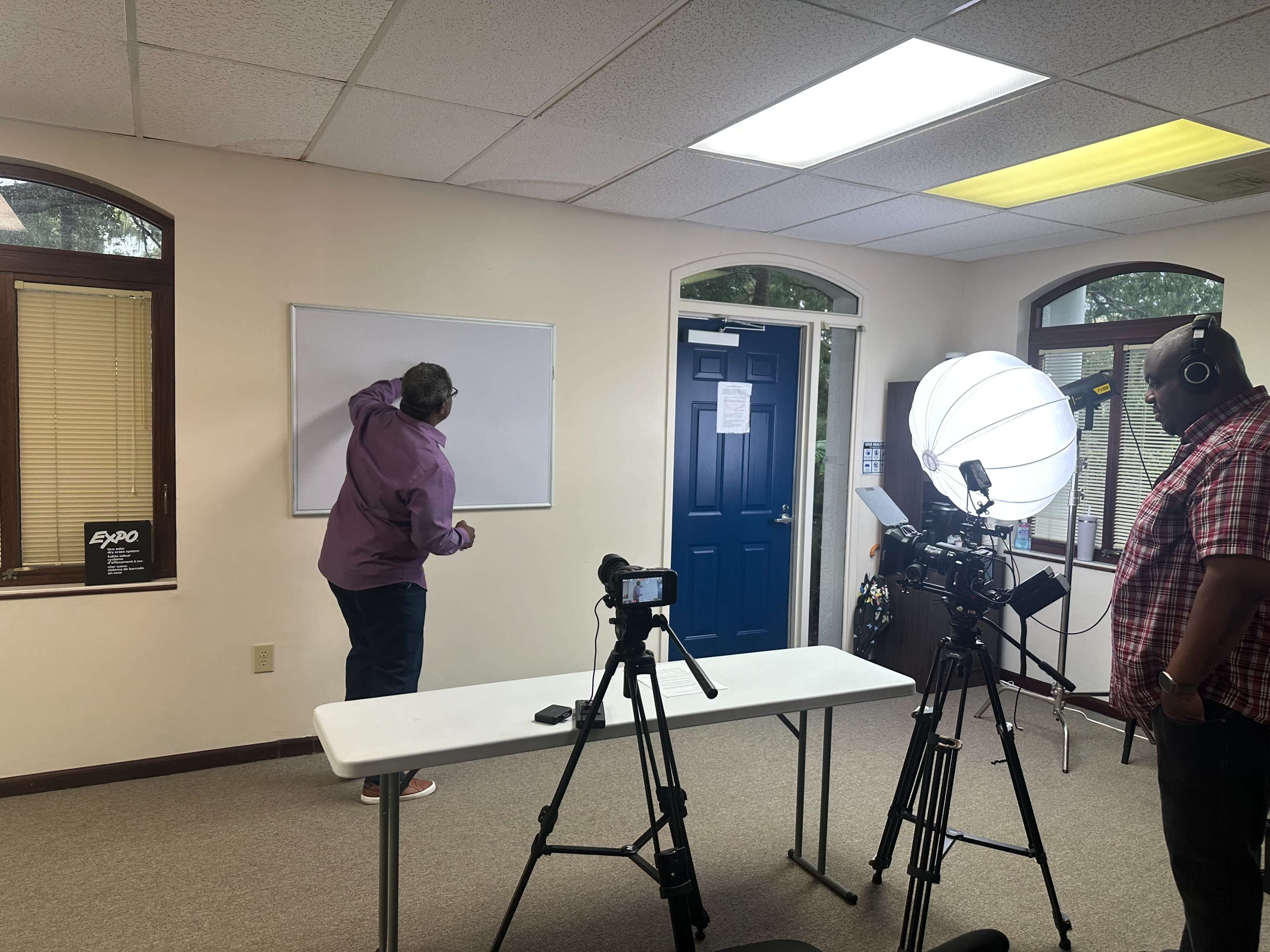 A person writes on a whiteboard while another person observes and prepares equipment in a room set up for video recording.