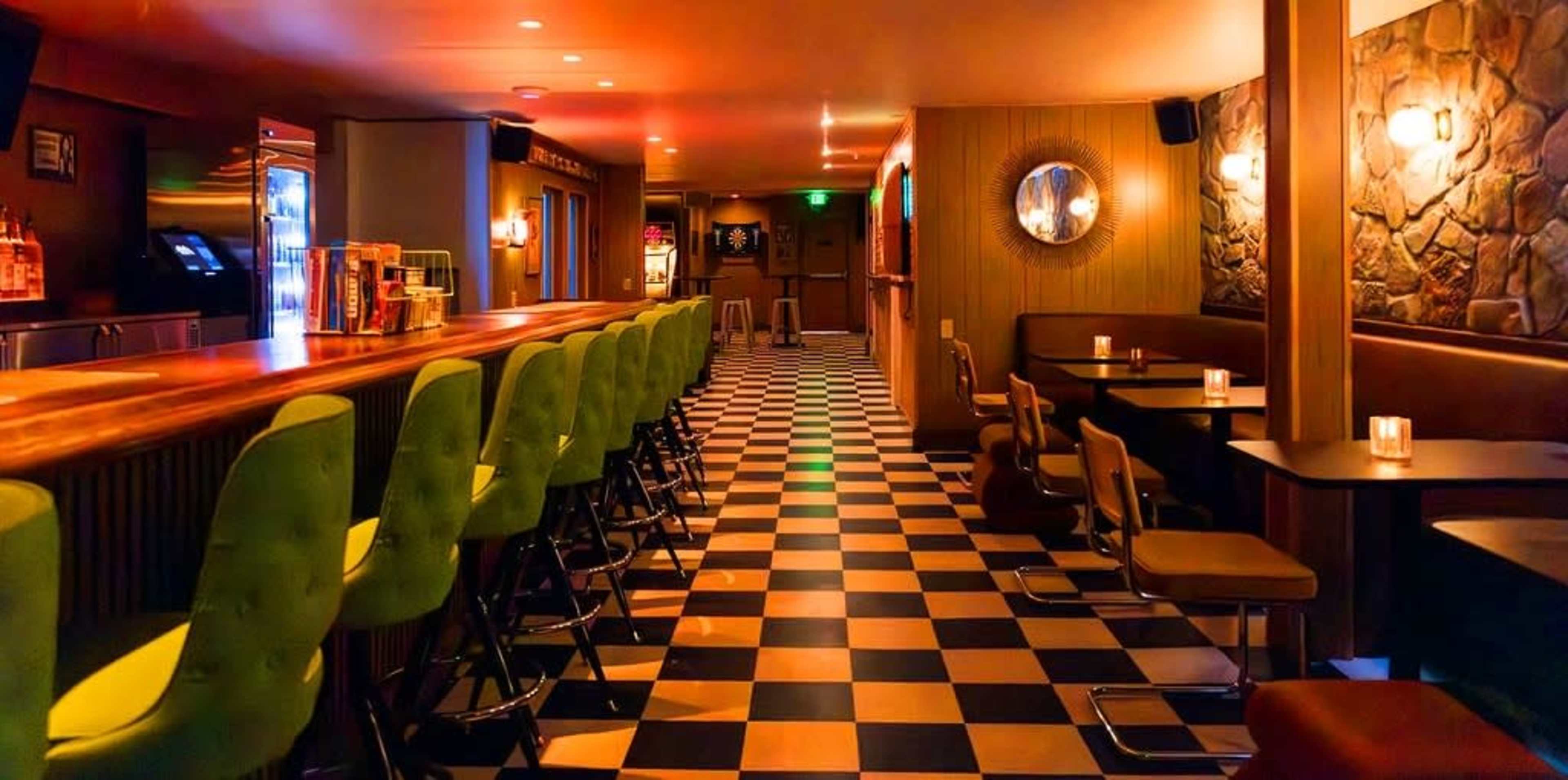 The image shows an empty bar with a long wooden counter, green high stools, and a checkered floor.