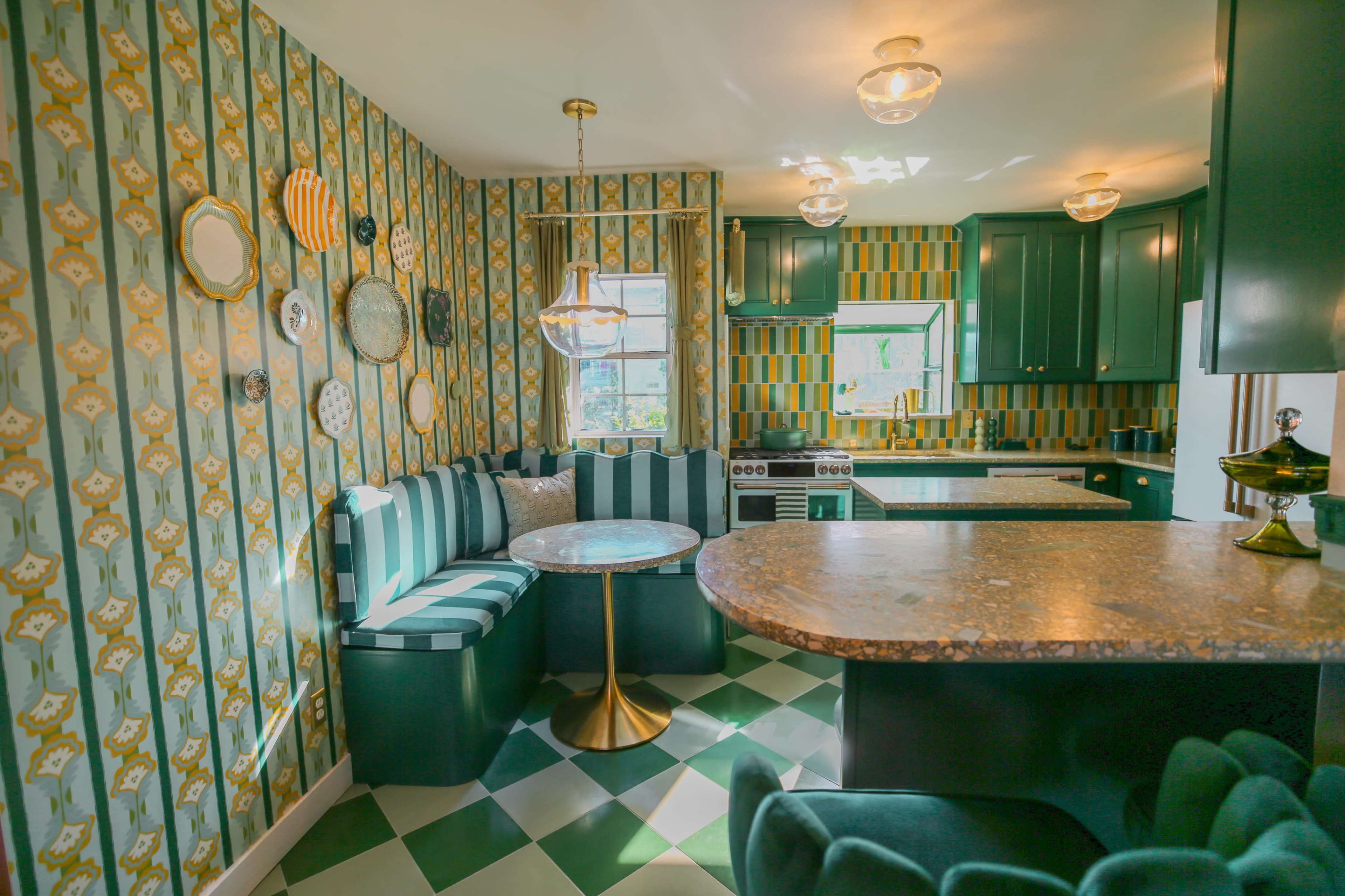 A compact kitchen features green cabinetry, striped seating, and patterned wallpaper, with a round table illuminated by pendant lights.
