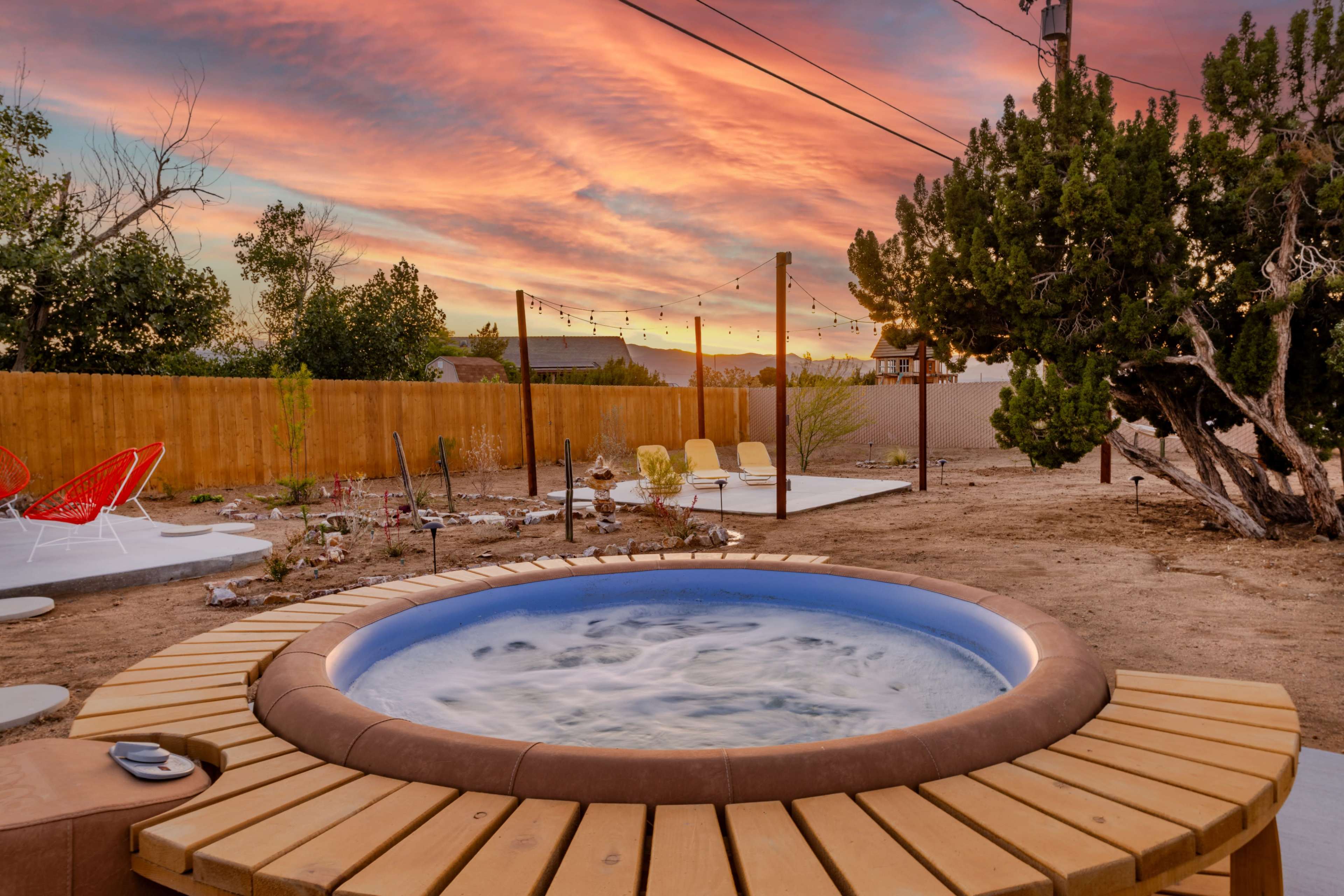 A hot tub with a wooden deck sits in a backyard at sunset, surrounded by trees and a wooden fence.