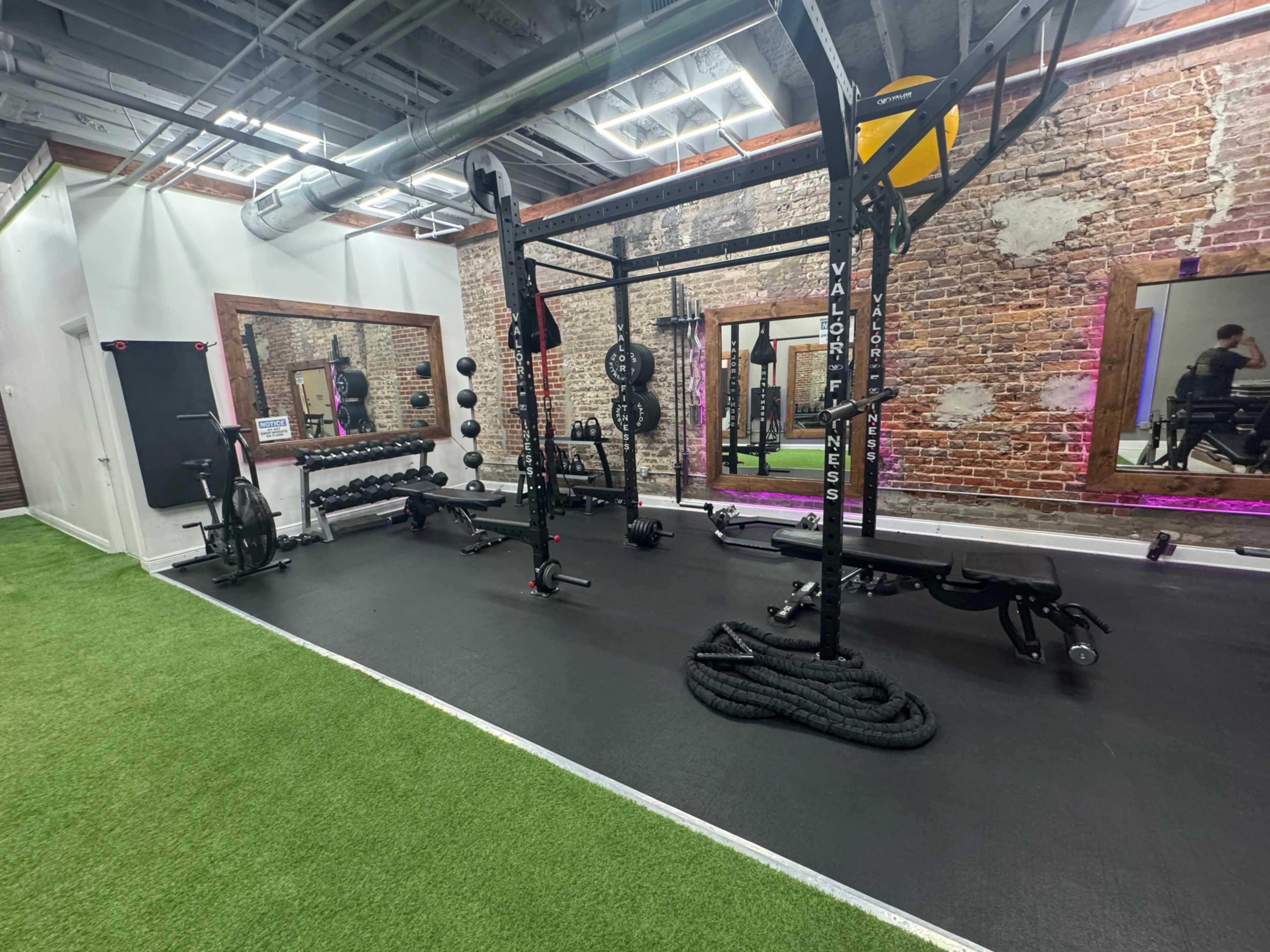 The image shows a modern gym featuring various exercise equipment, including weightlifting racks, dumbbells, and battle ropes, with a brick wall backdrop and green turf flooring.
