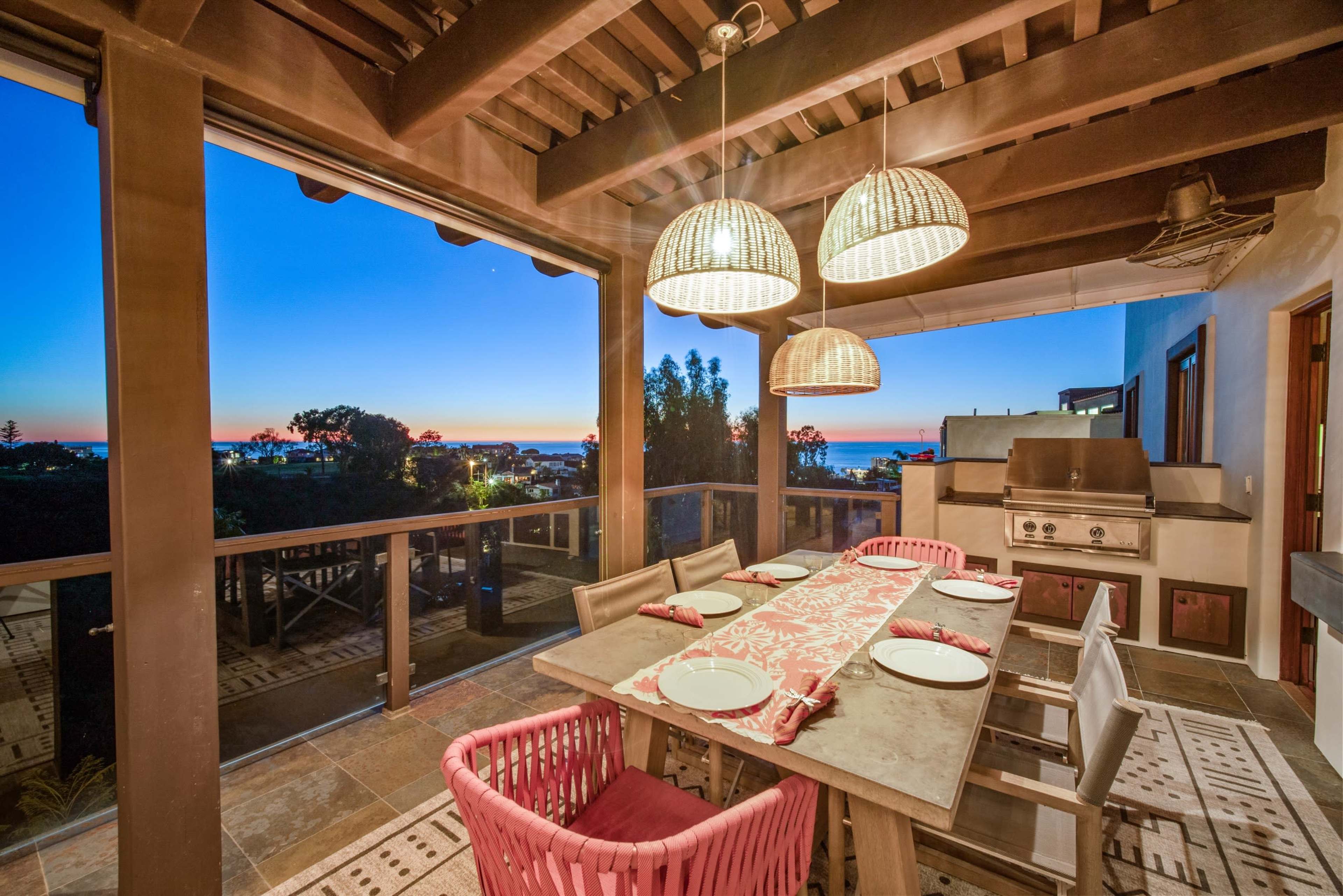 The image shows a dining area on a patio with a table set for a meal, overlooking a sunset view with ocean and landscape visible in the background.