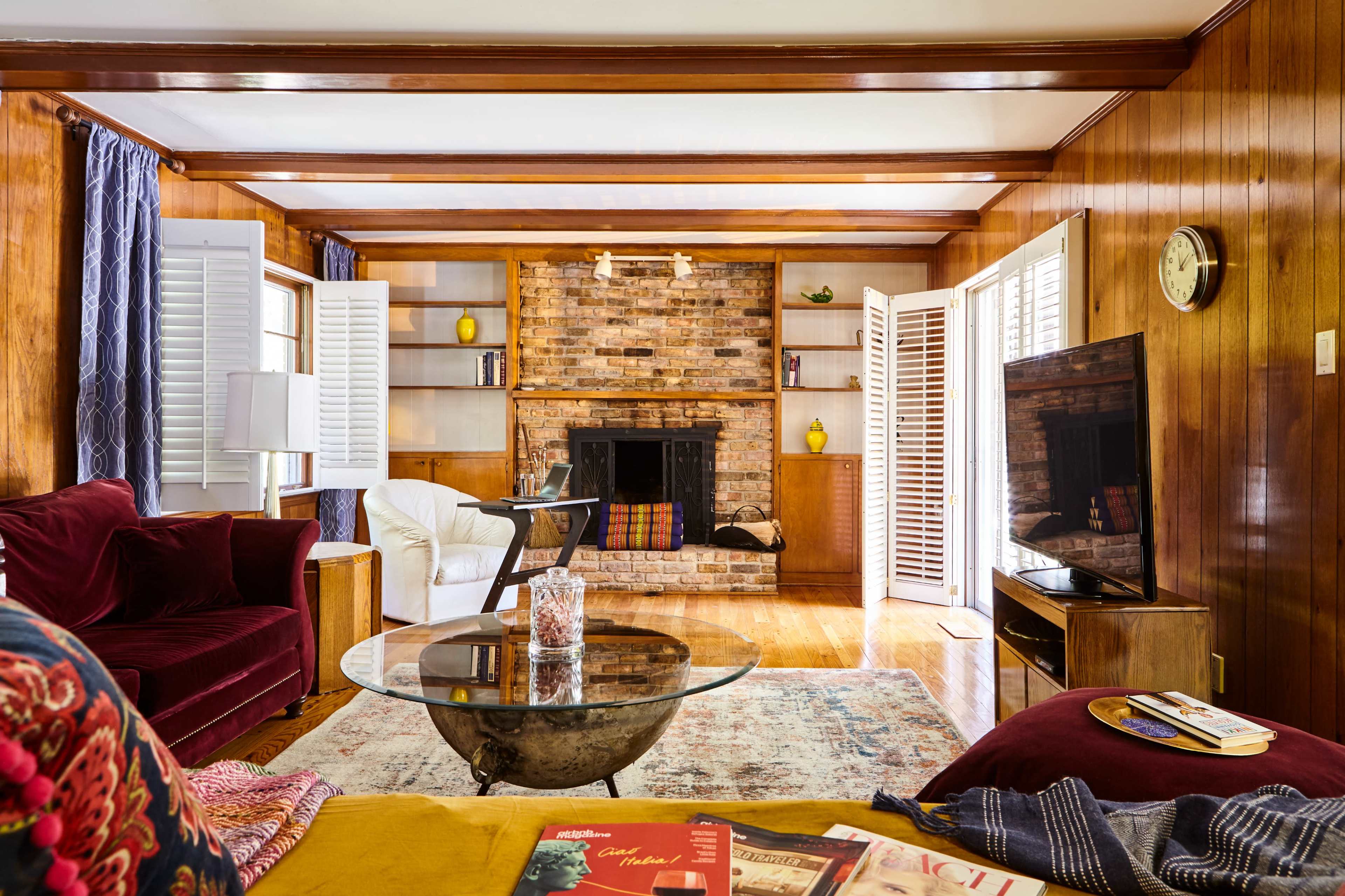 The living room features a brick fireplace, wooden paneling, and a combination of seating areas, including a red couch and a white armchair.