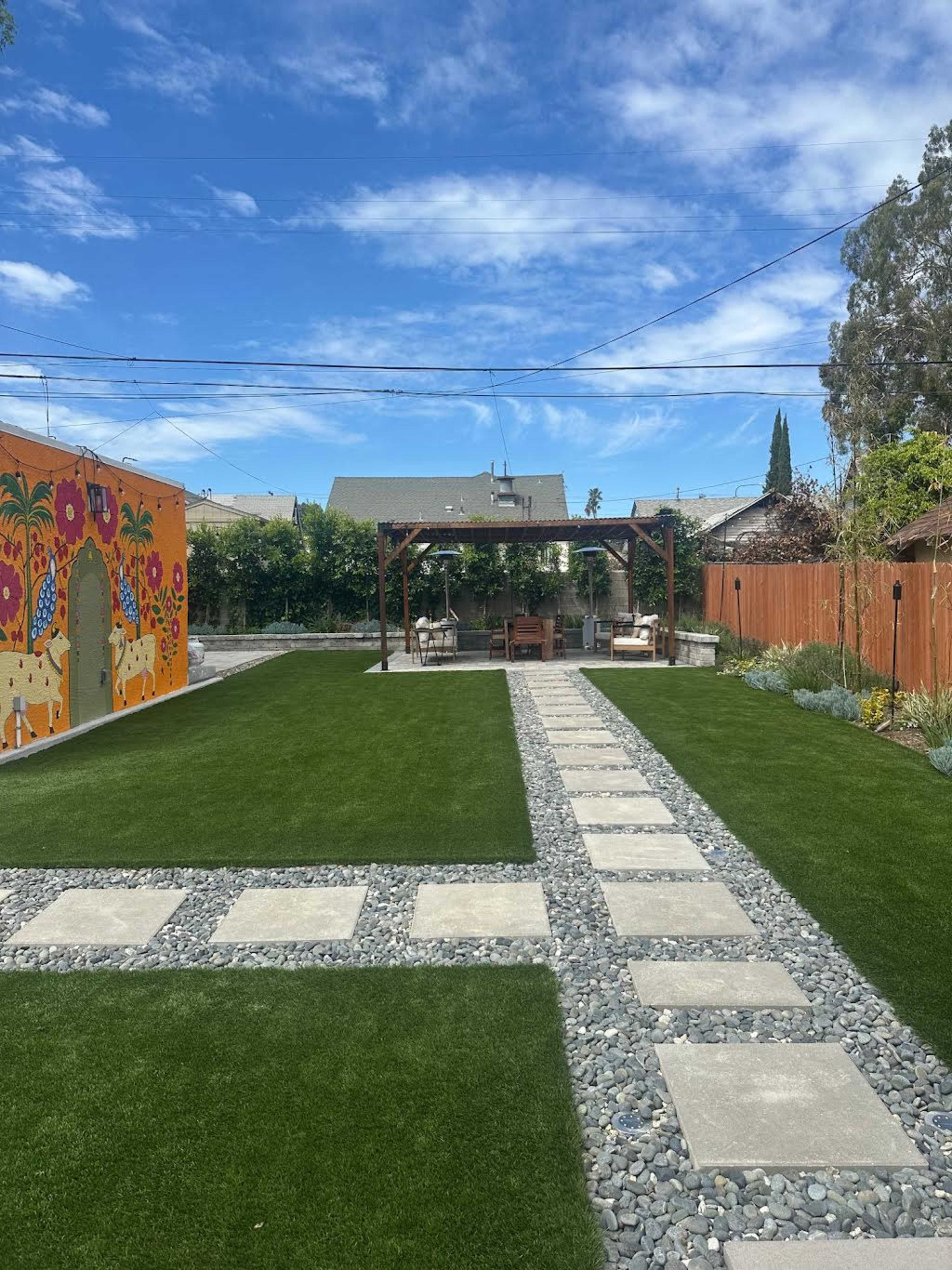 The scene features a landscaped backyard with a gravel pathway, concrete stepping stones, and a vibrant mural on one wall, surrounded by green grass and a gazebo.