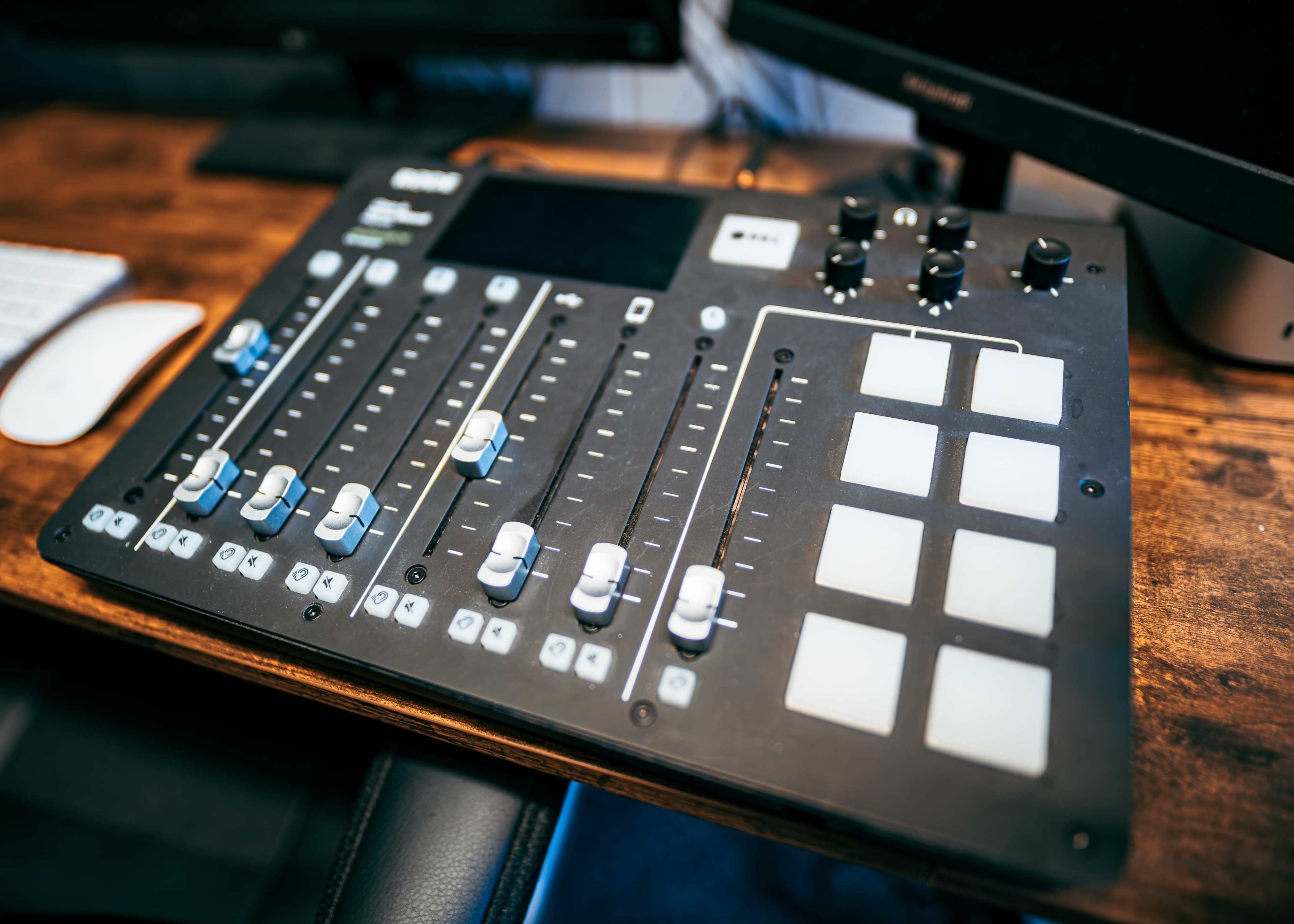 The image shows a professional audio mixing console with faders, knobs, and a grid of buttons on a wooden surface.