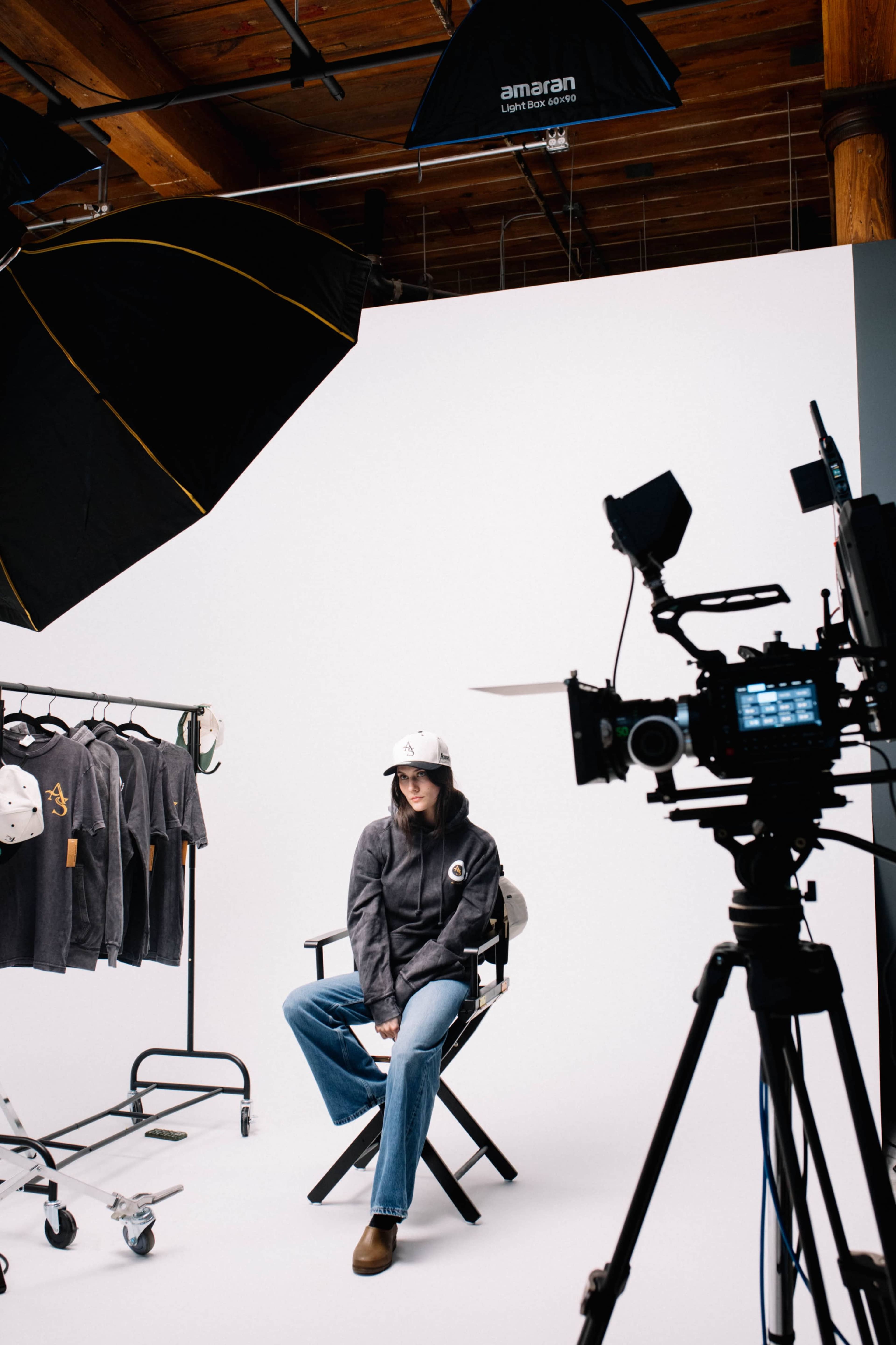 A model sits on a director's chair in a studio with clothing on a rack and camera equipment nearby.