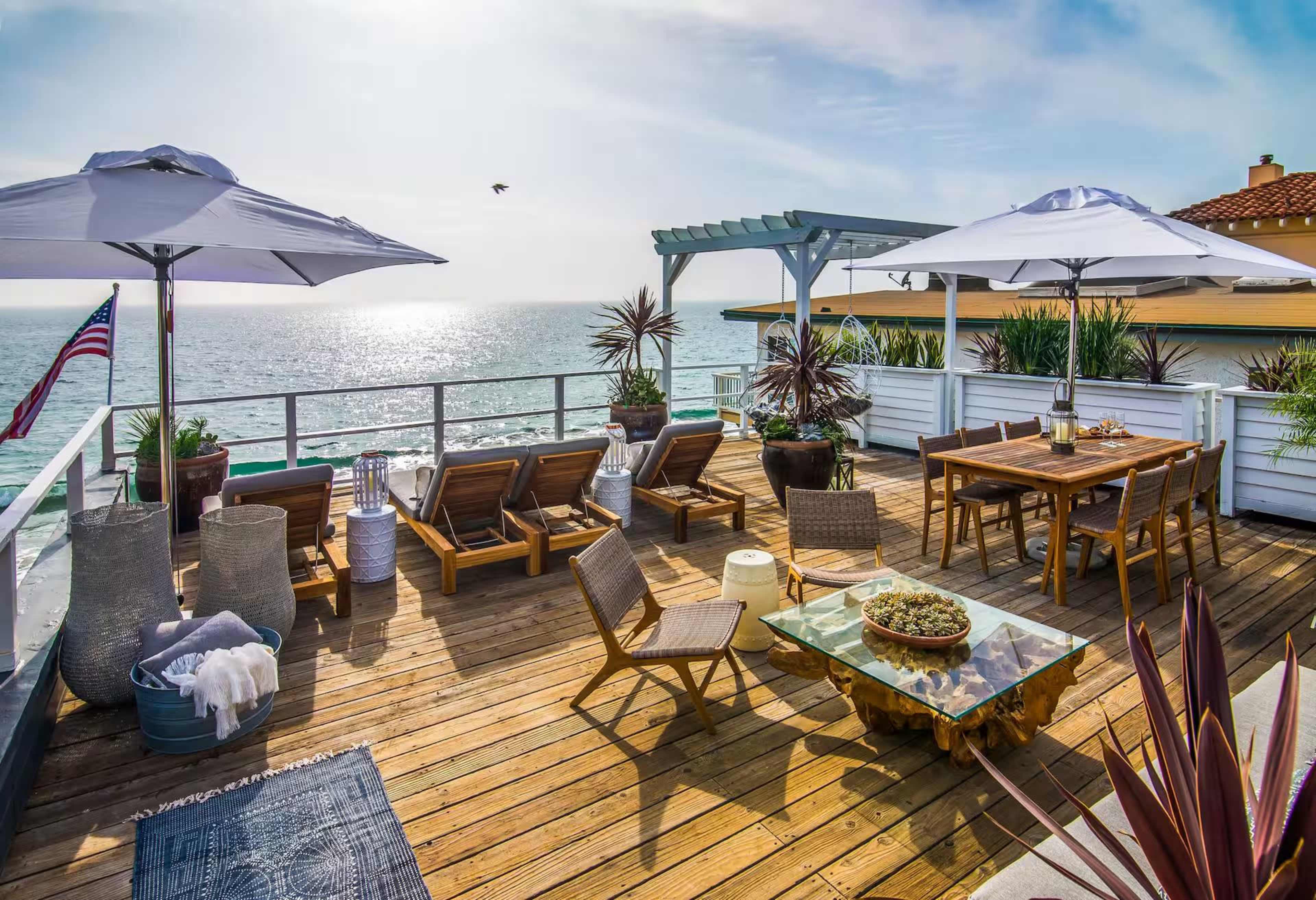 A wooden deck features lounge chairs, a dining table, and umbrellas overlooking the ocean at sunset.