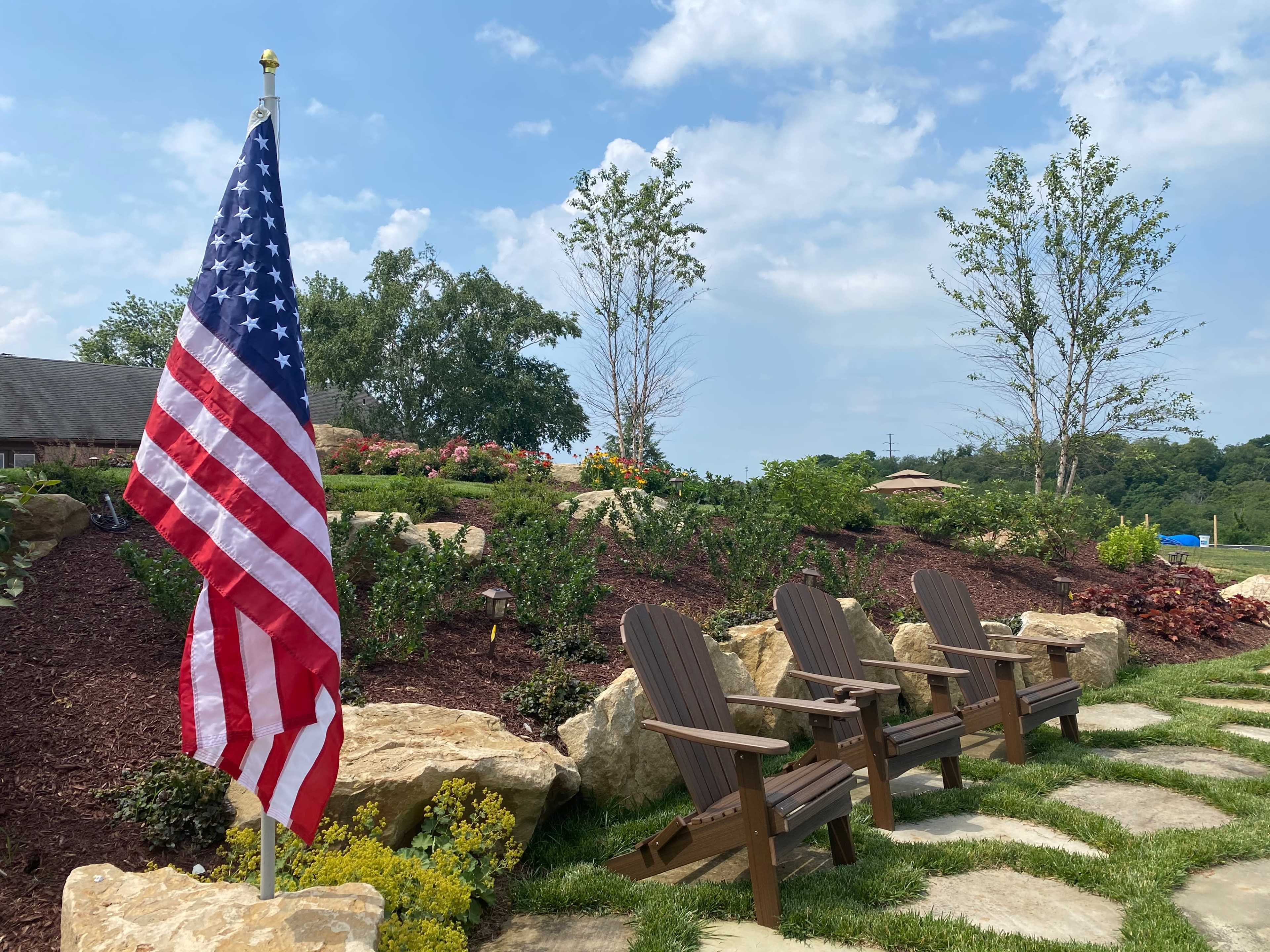 A U.S. flag stands next to a pathway lined with stone, leading to three wooden chairs amidst landscaped greenery and flowers.
