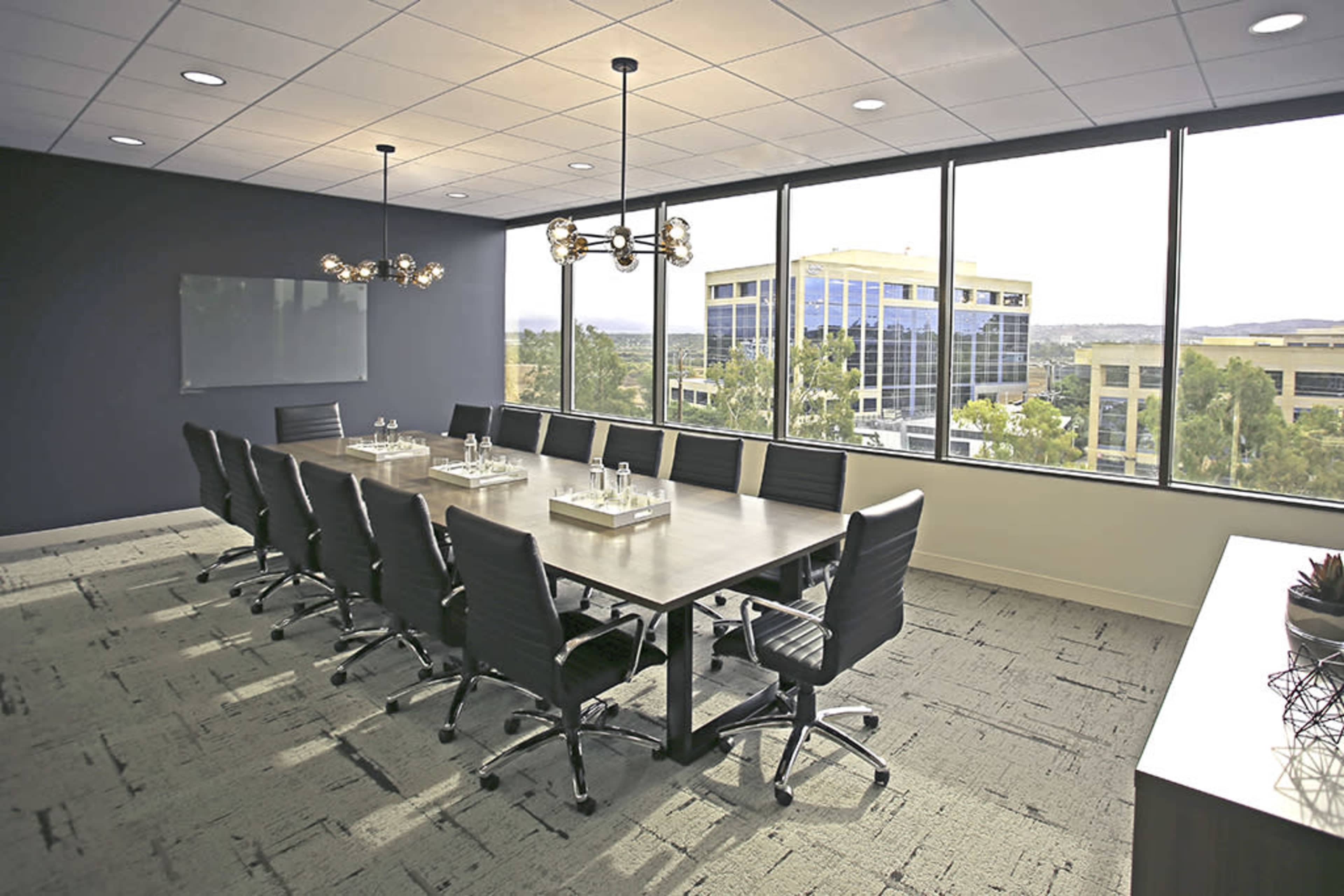 A modern conference room features a large table surrounded by black chairs, with windows showcasing an outdoor view of office buildings and greenery.