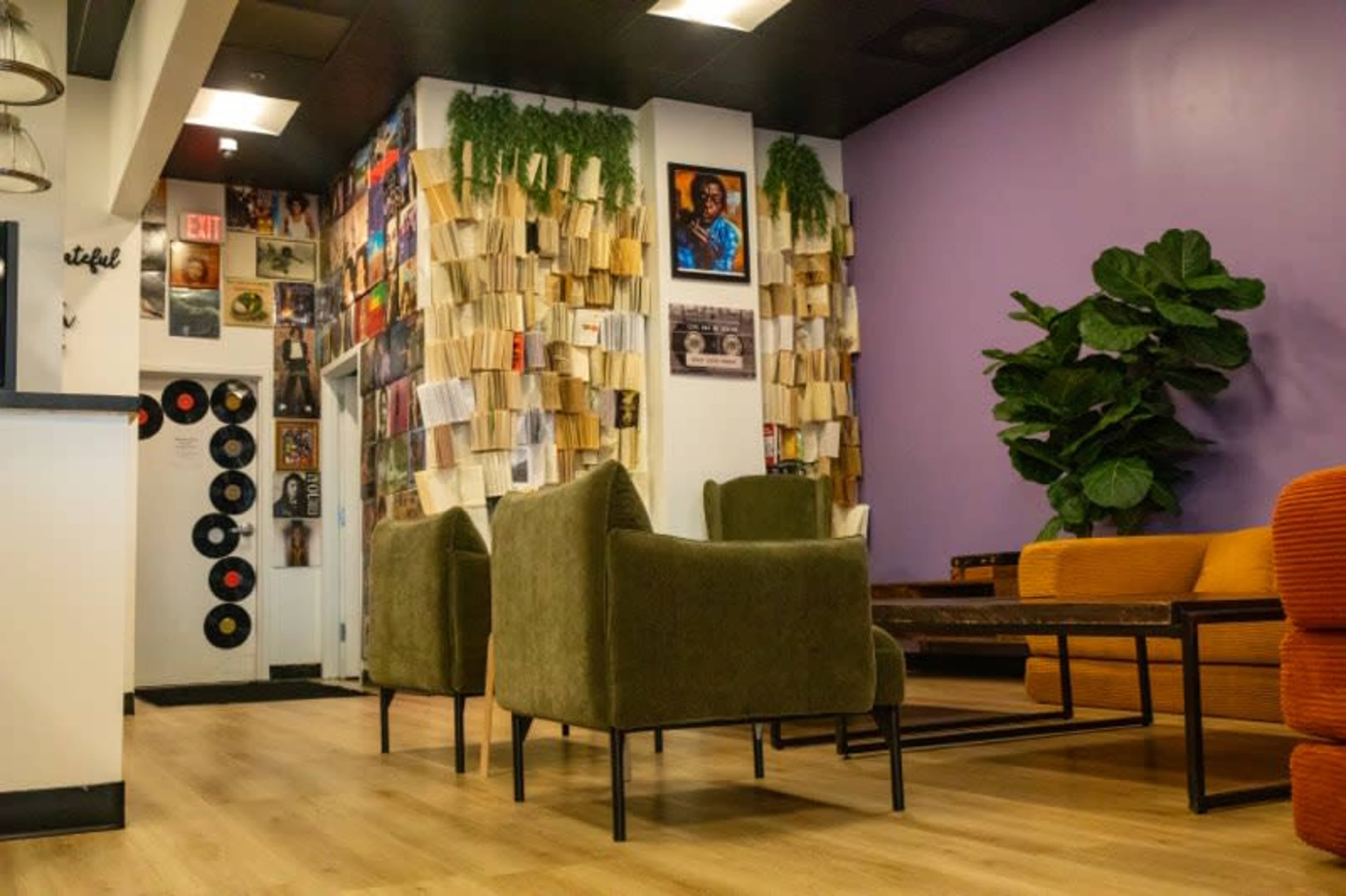 The image shows a modern lounge area with a purple wall, bookshelves filled with records, two green armchairs, and a large potted plant.