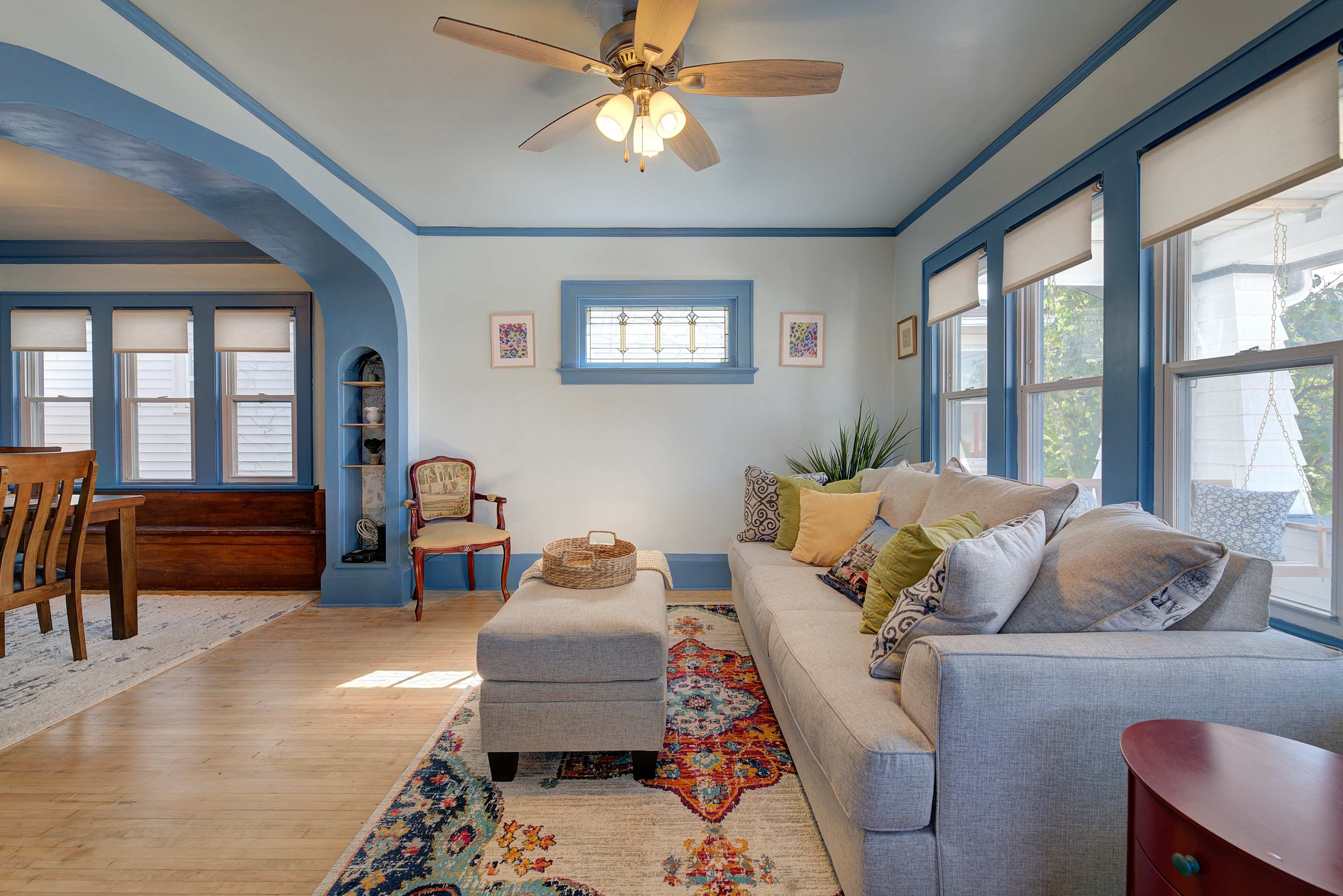 A living room with a light blue color scheme, featuring a beige sofa, an ottoman, decorative pillows, and large windows that allow natural light to illuminate the space.