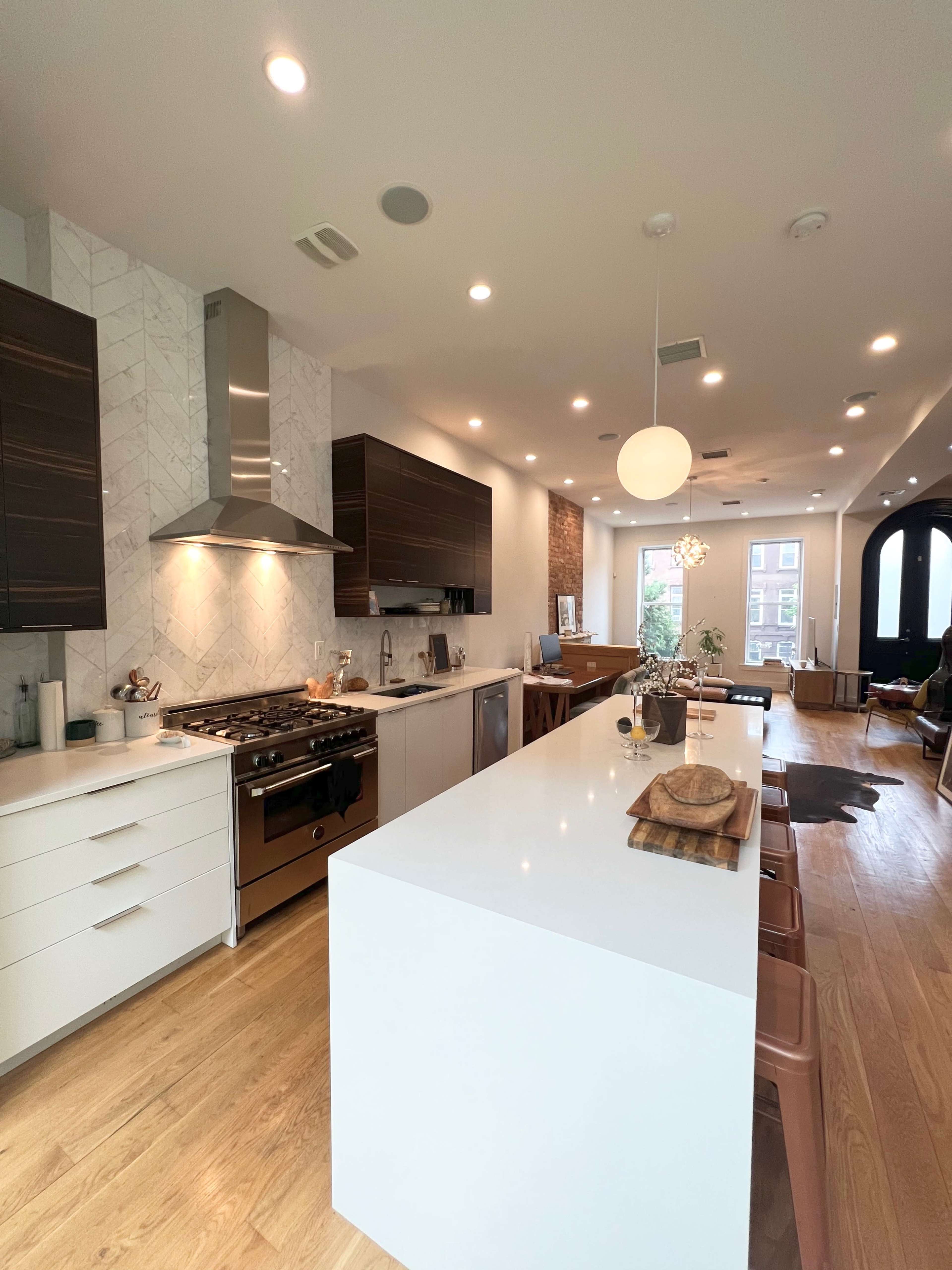 The image shows a modern kitchen featuring a large island, stainless steel appliances, and ample cabinetry, with a dining area visible in the background.