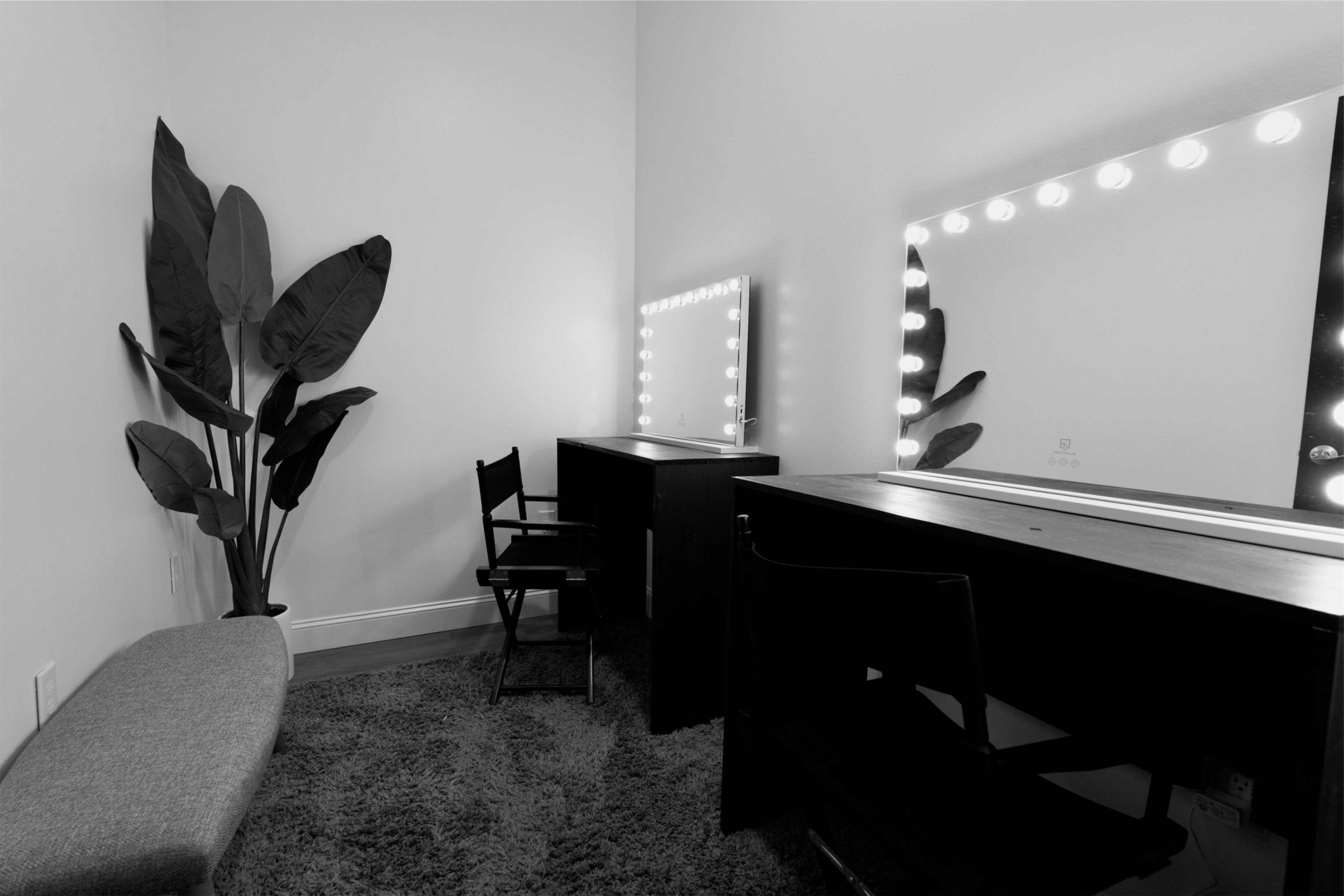 A small, minimalist makeup area with two mirrors framed by light bulbs, a gray bench, a potted plant, and two black chairs.