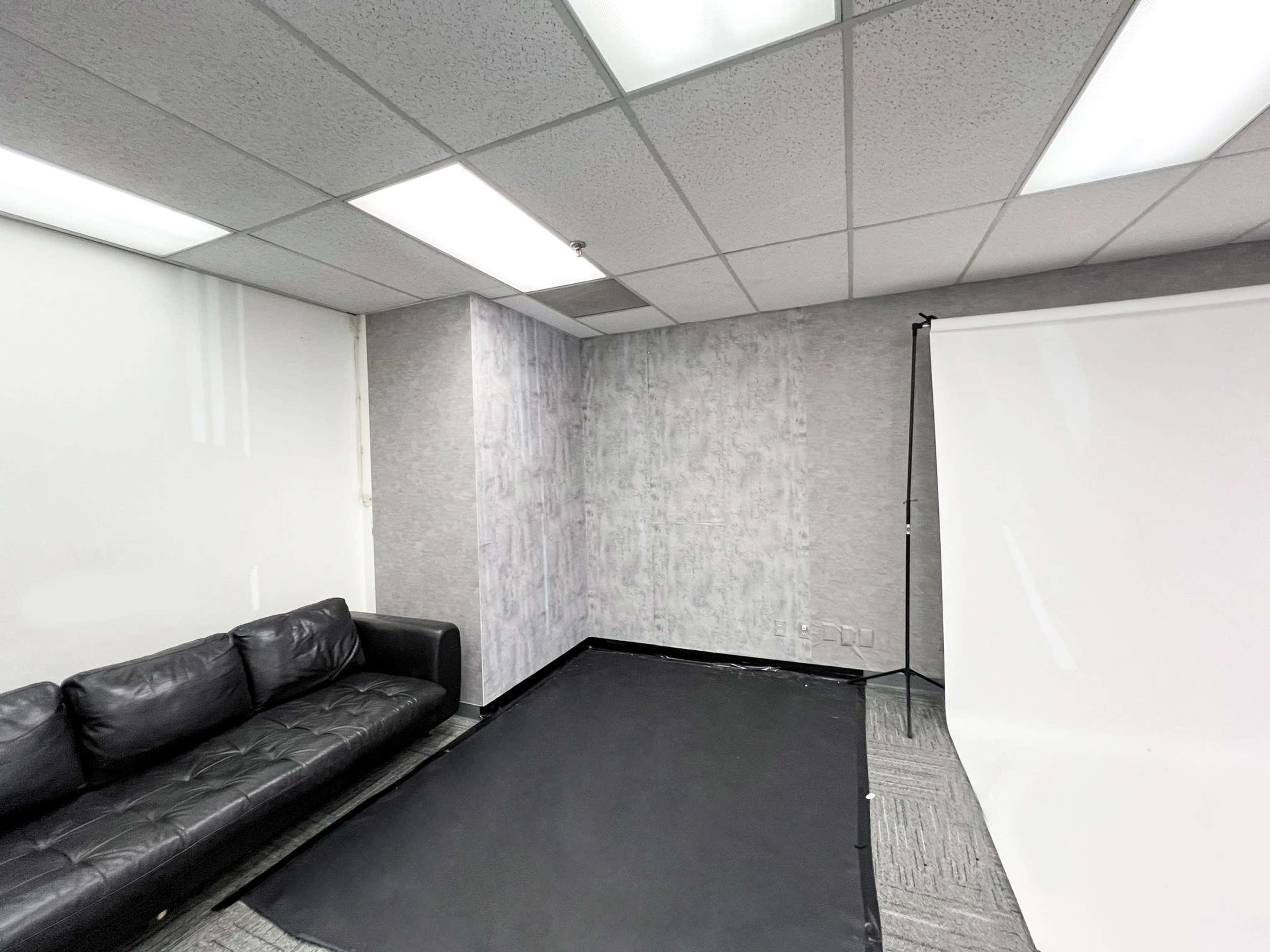 A small room featuring a black floor mat, a gray textured wall, a white backdrop on a stand, and a black leather sofa.