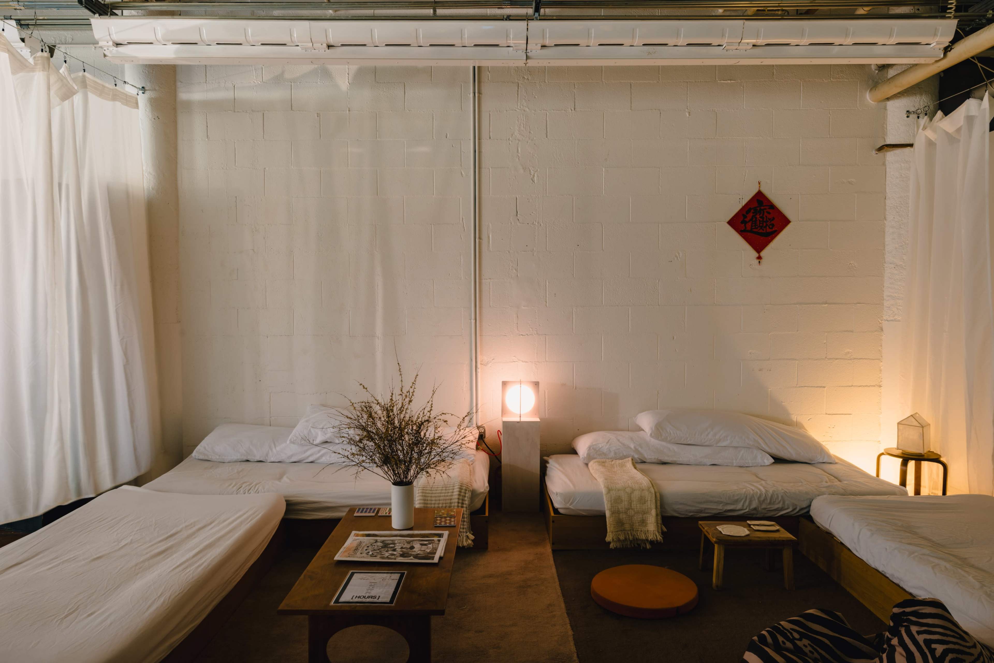 The image shows a minimalist bedroom setup with three beds, soft lighting, and white curtains dividing the space.