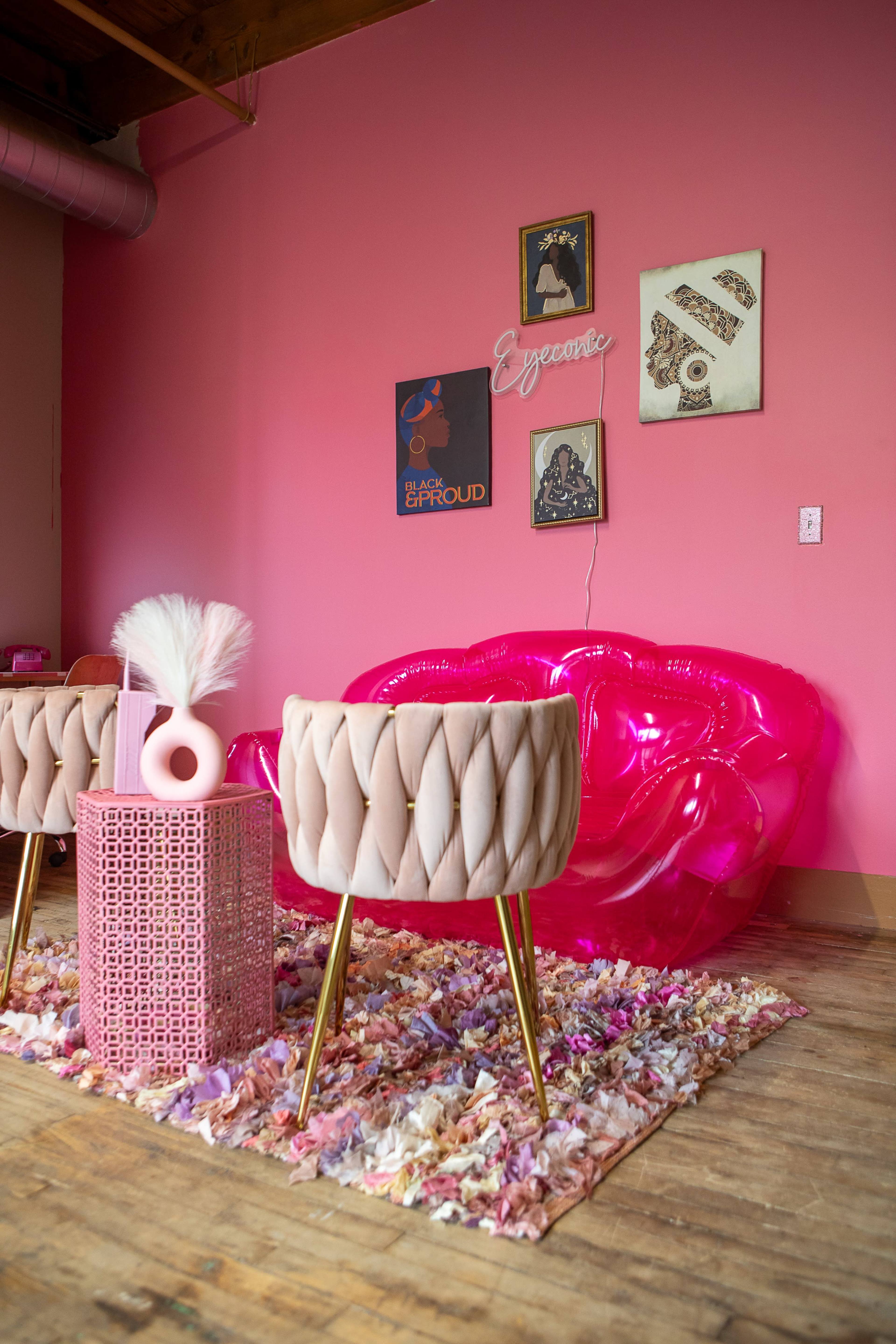 A pink-themed lounge area features a transparent lip-shaped sofa, two upholstered chairs, a decorative rug, and framed art on the walls.